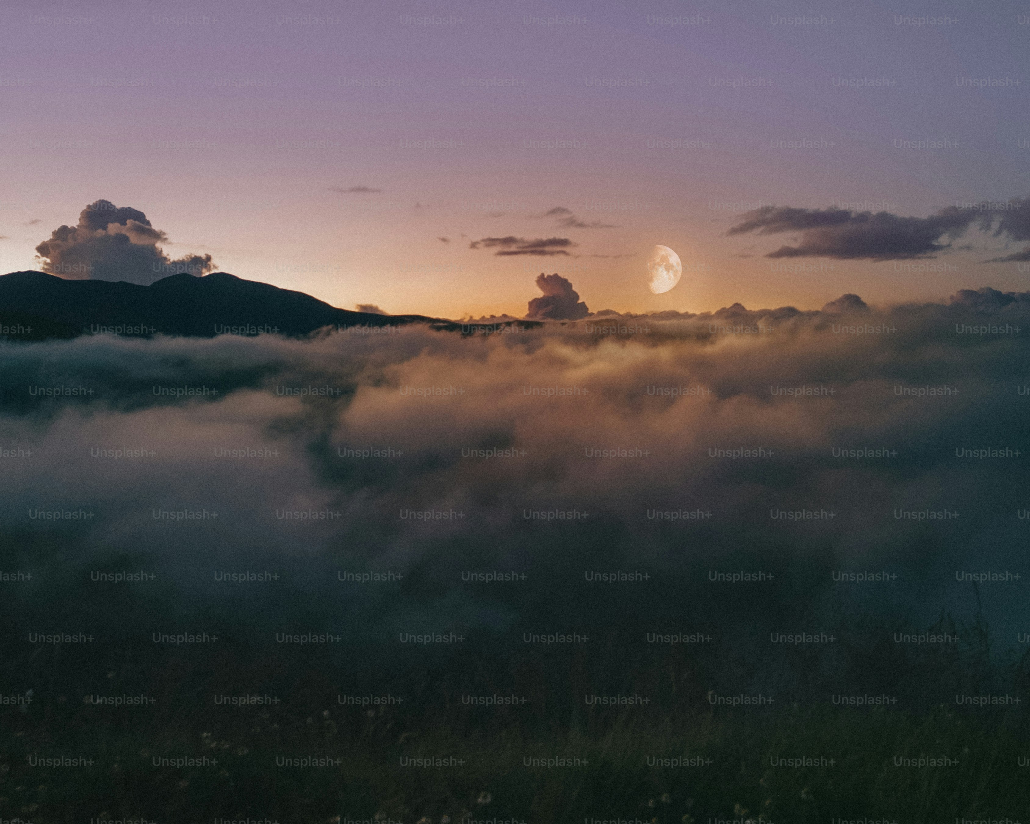 Crescent moon above clouds at sunset over mountains.