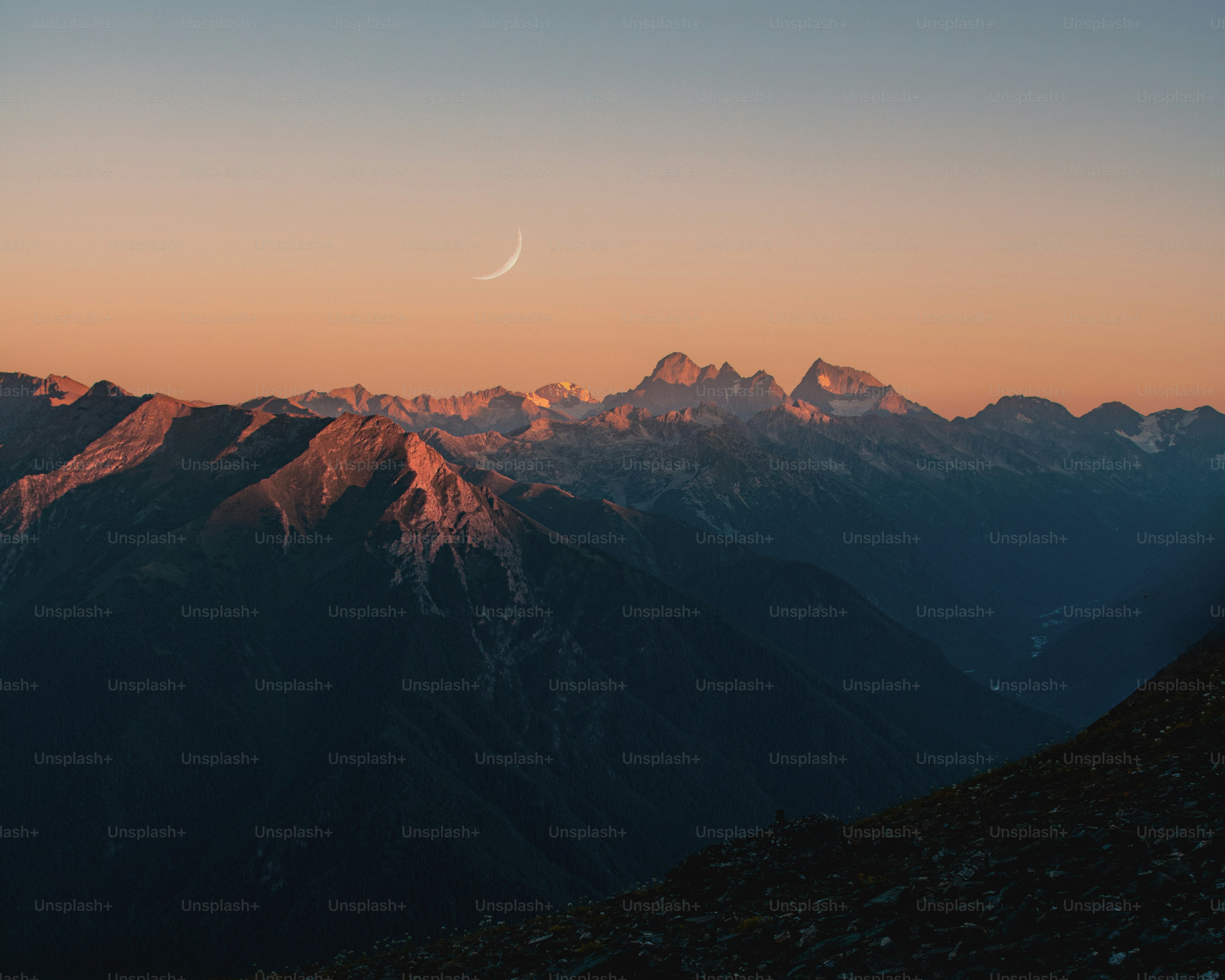Crescent moon over sunlit mountain peaks at dawn