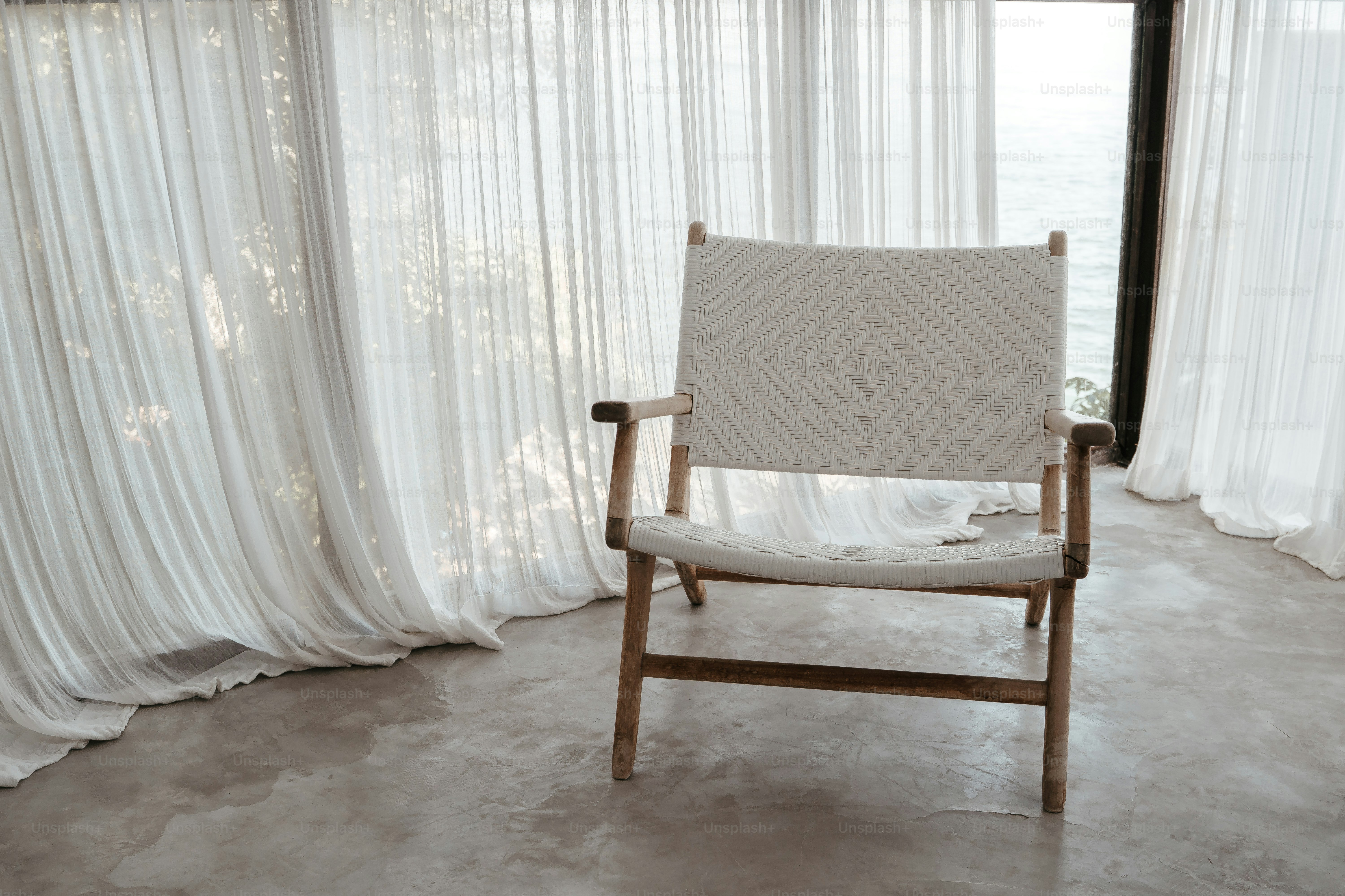 A minimalist chair sits near white curtains and a window.