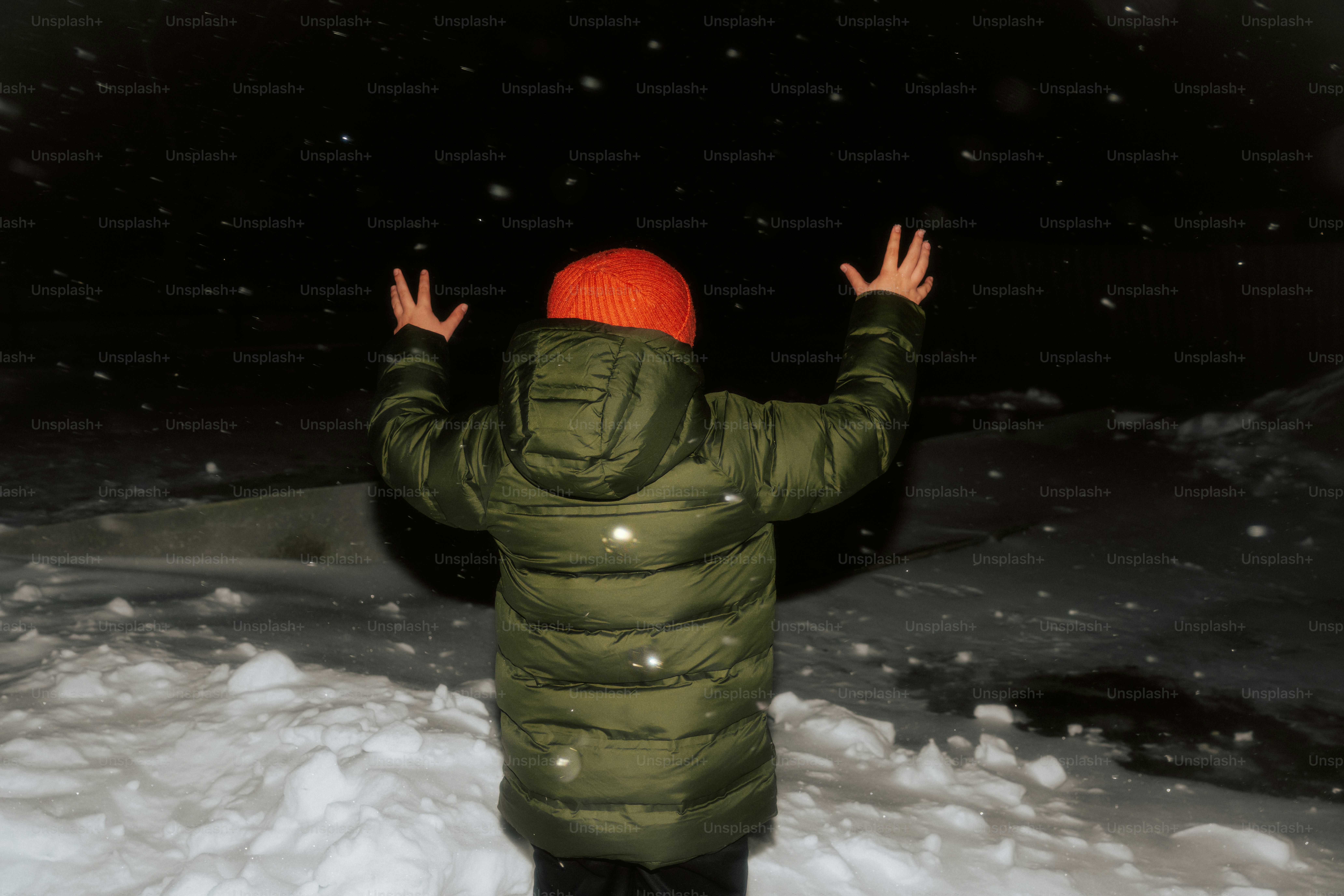 A child in a puffer coat catches snowflakes at night.