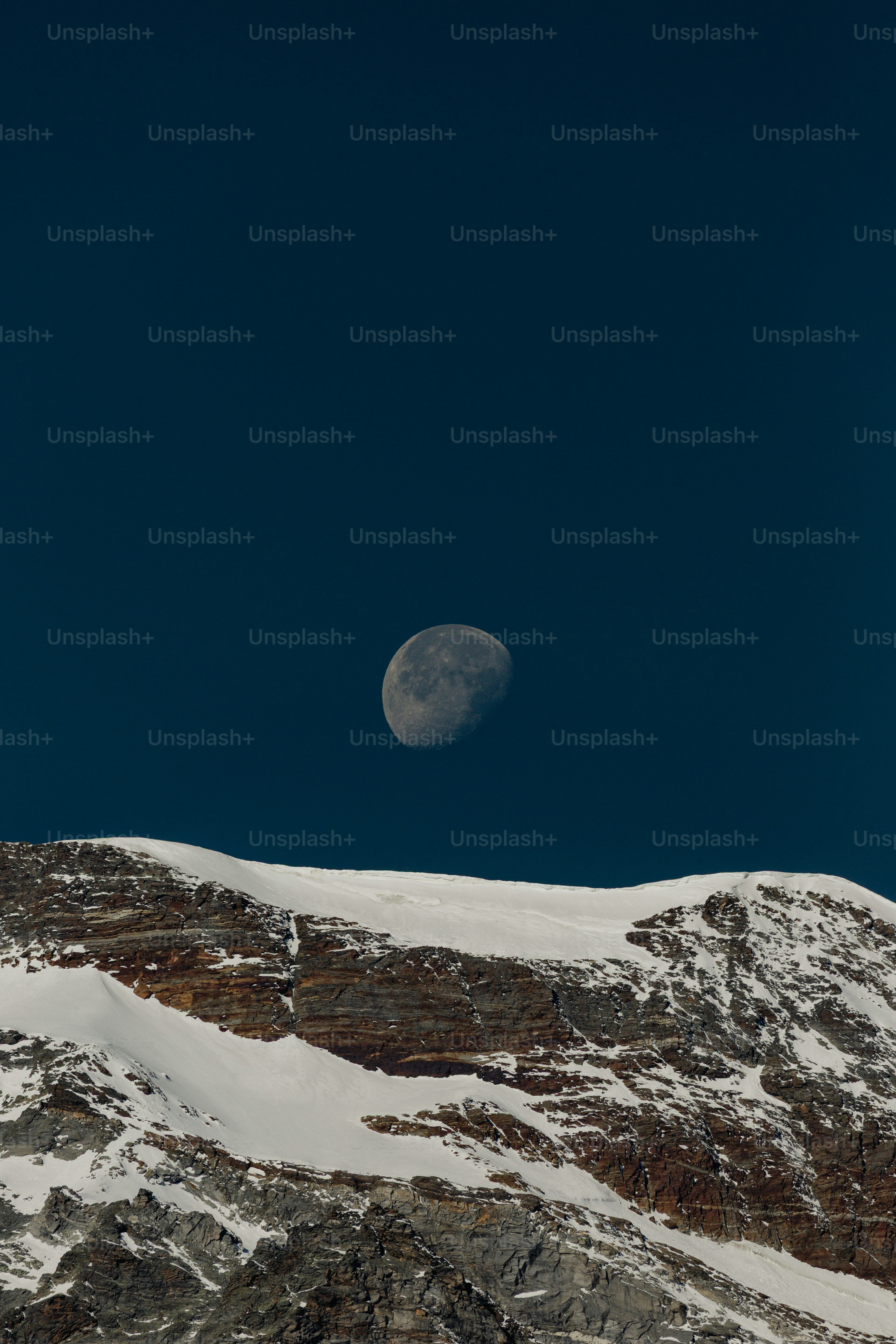 Half moon above snow-capped mountains under a clear blue sky