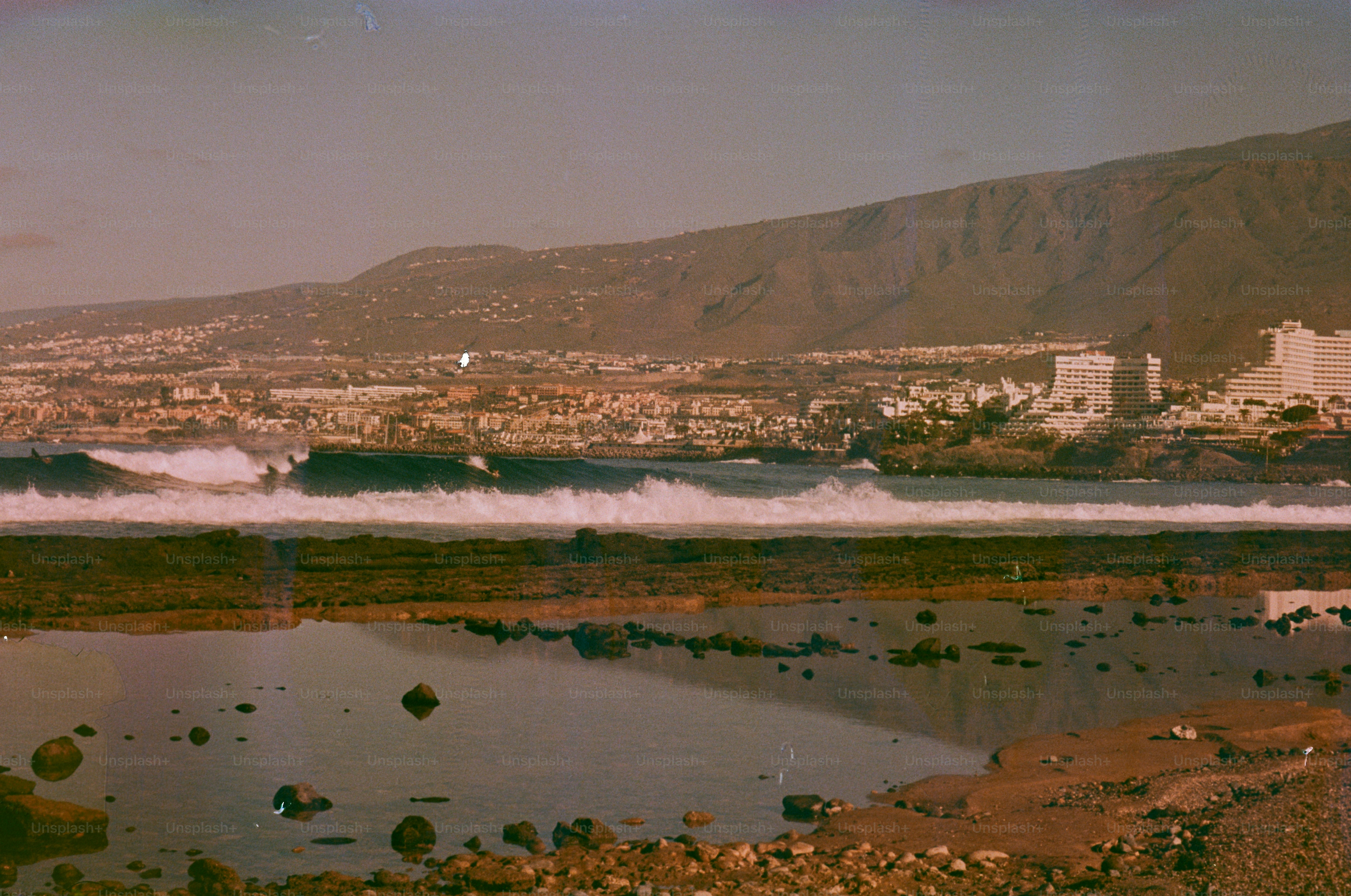 Waves crash near a coastal city with mountains behind.