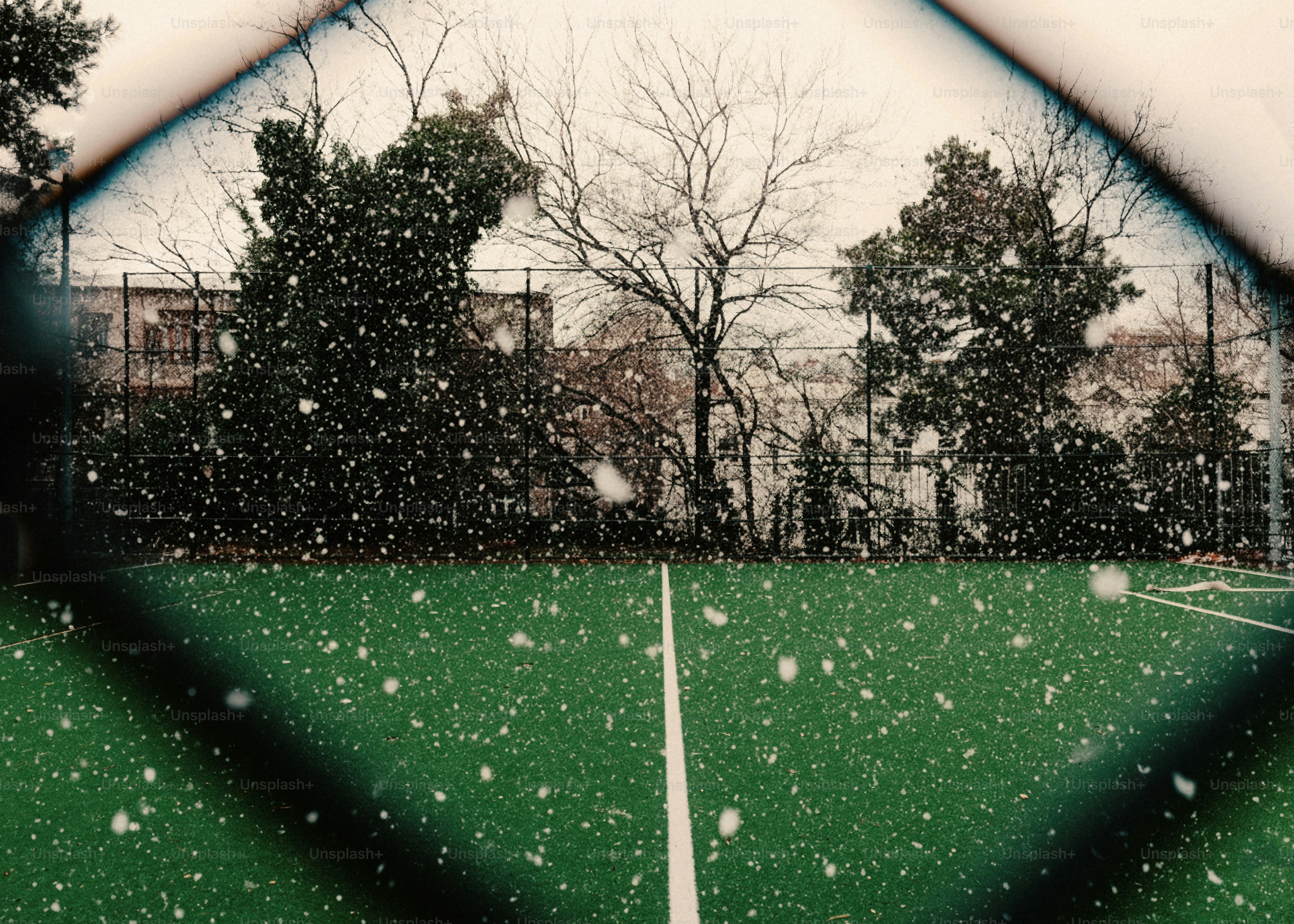 Snow falling on a green field through a fence.