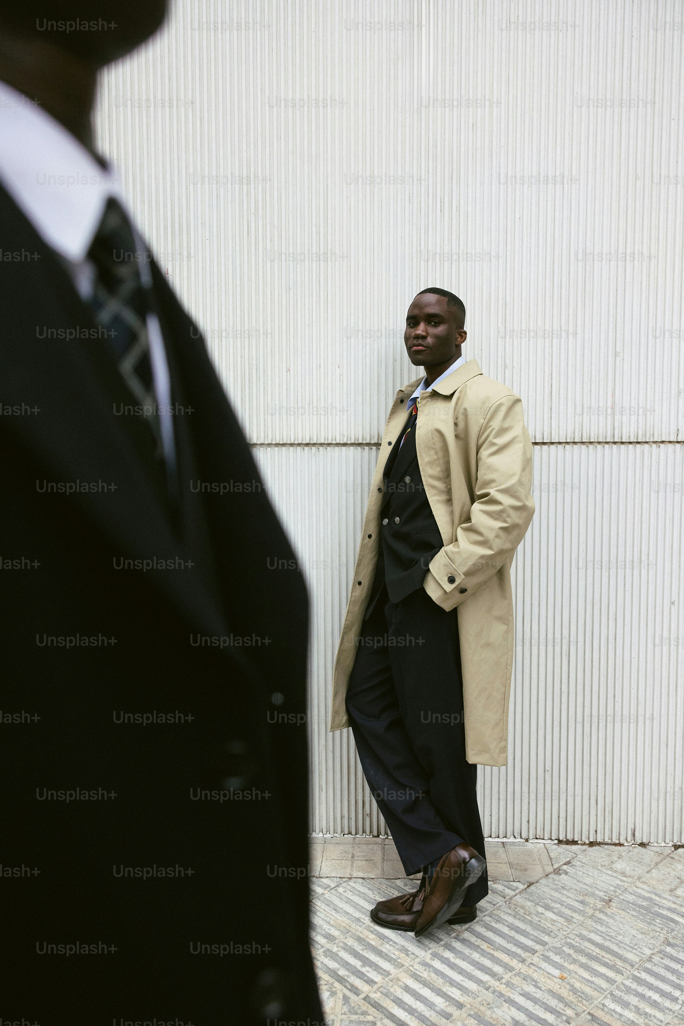 Two men in suits stand against a textured wall.