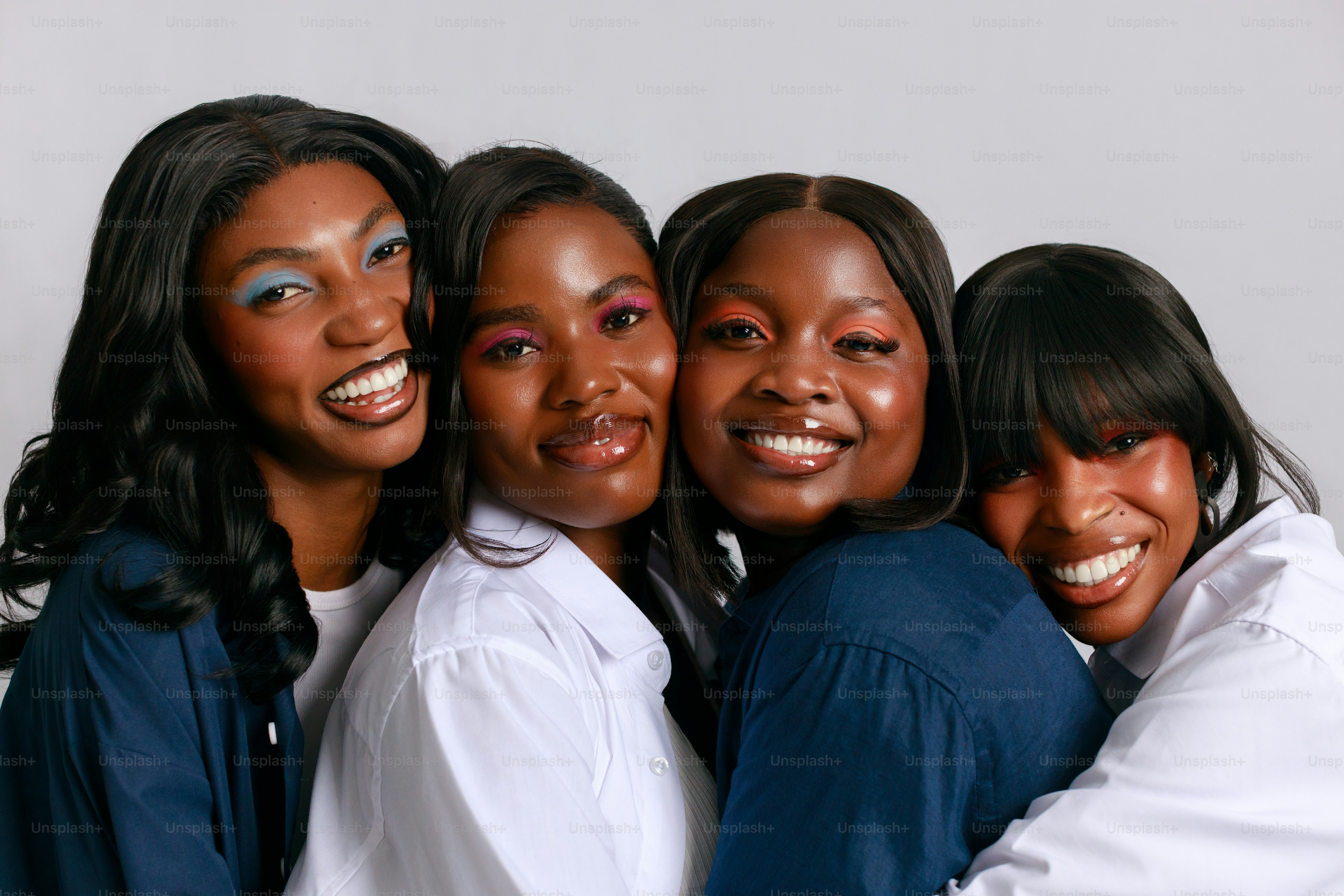 Four smiling women with vibrant makeup embrace closely.