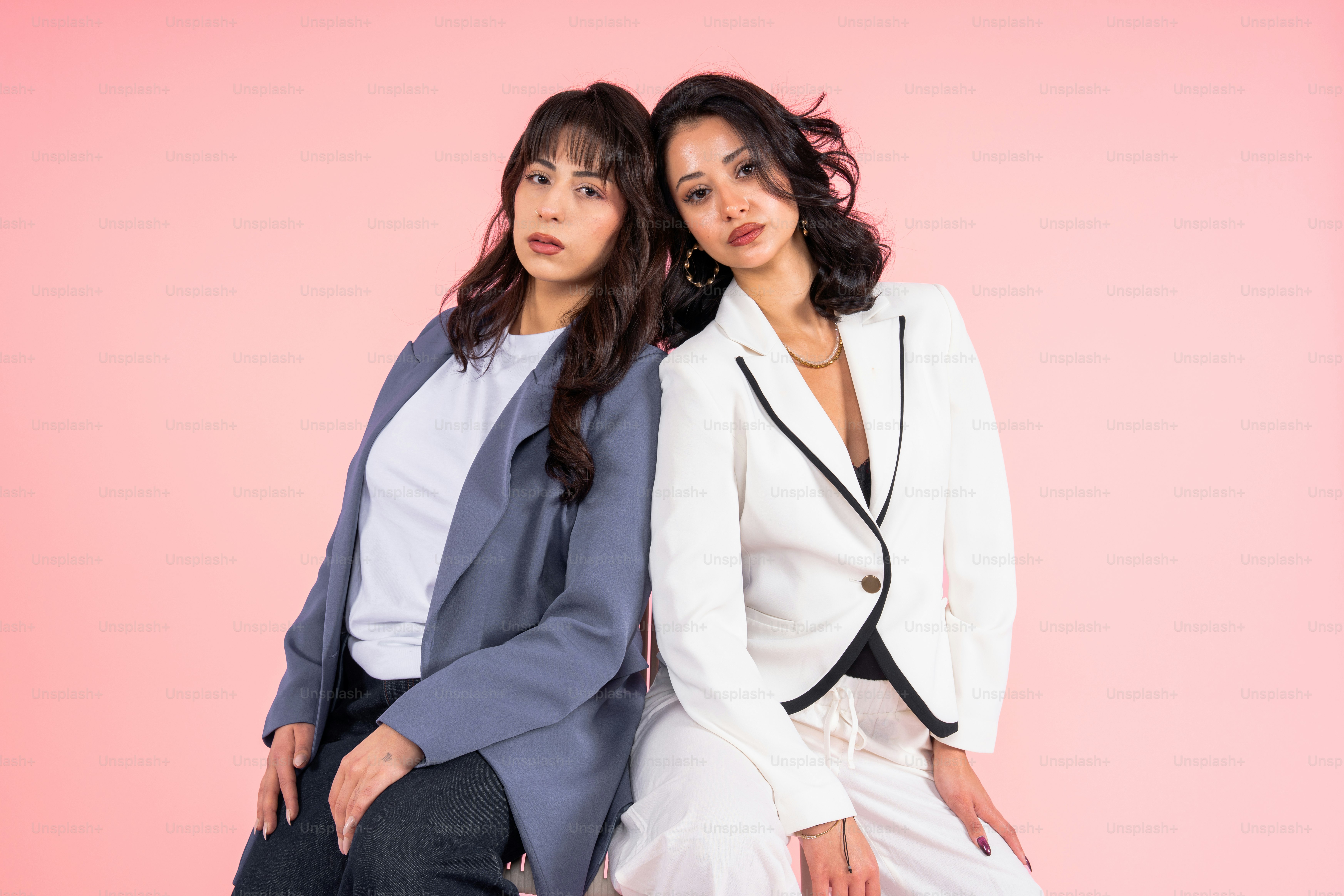 Two women in suits posing against a pink background.