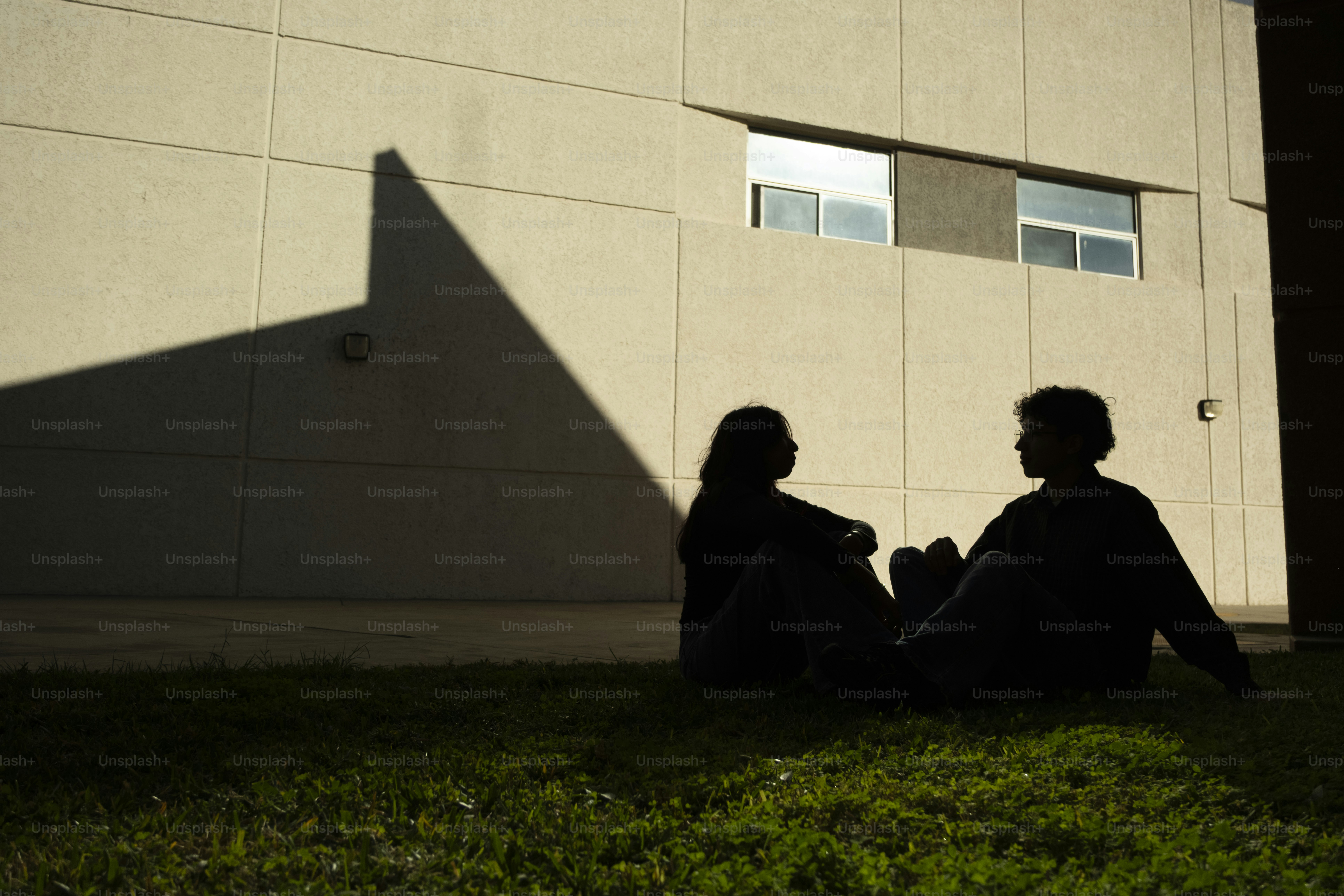 Two silhouetted figures sit on grass near building