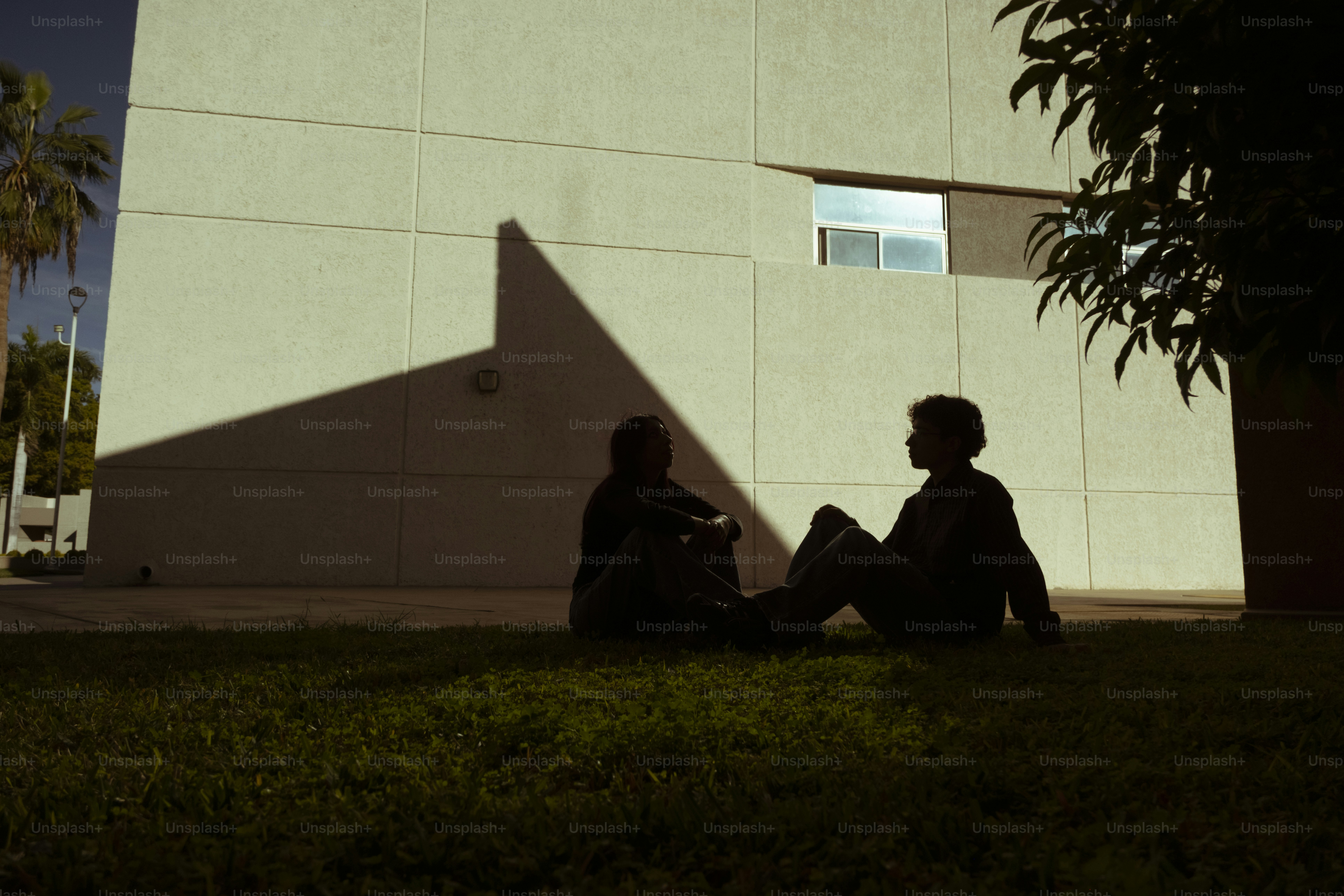 Two silhouetted figures sit on grass near a building.