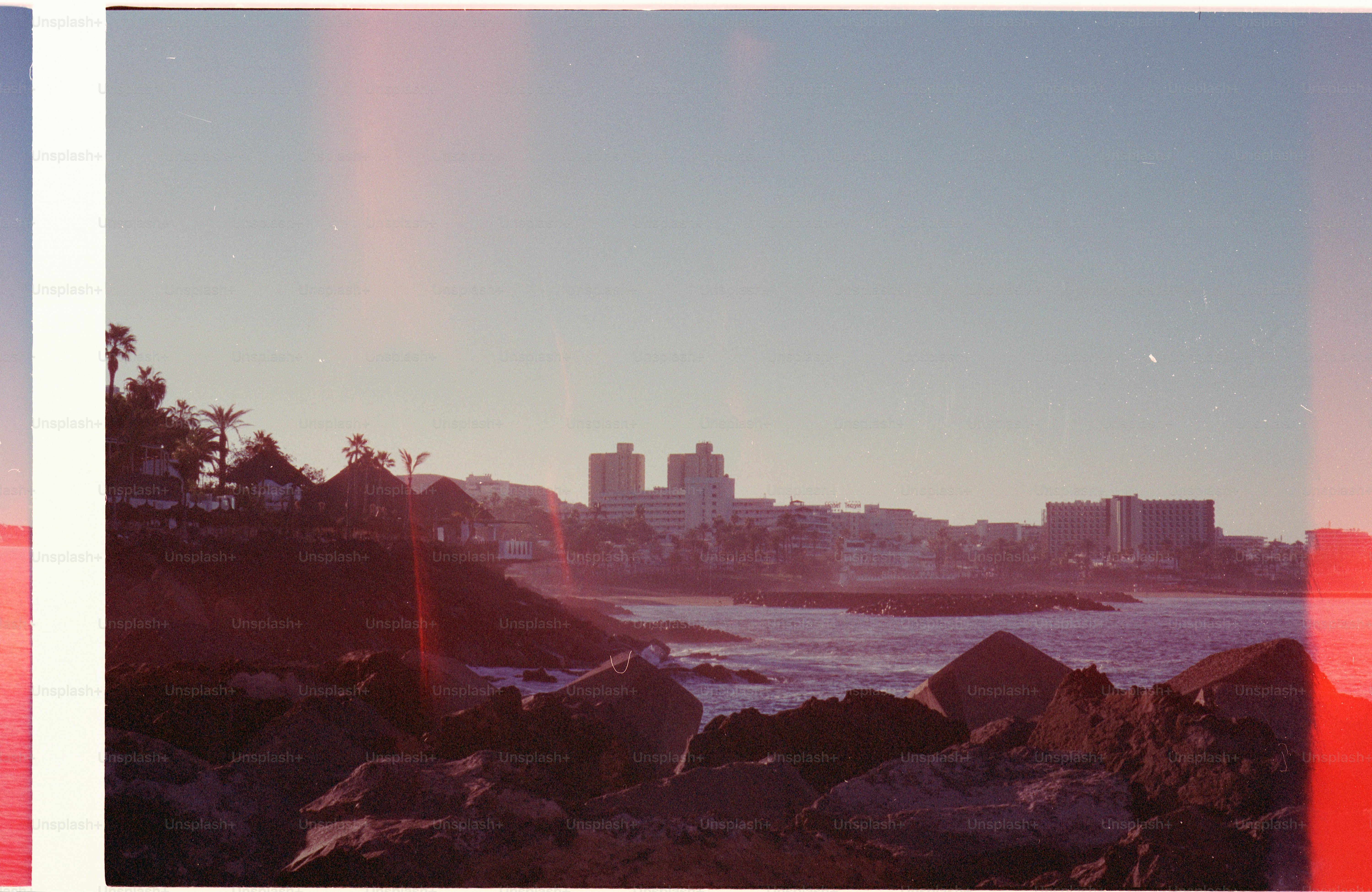 Coastal city skyline at sunset with rocky shore.