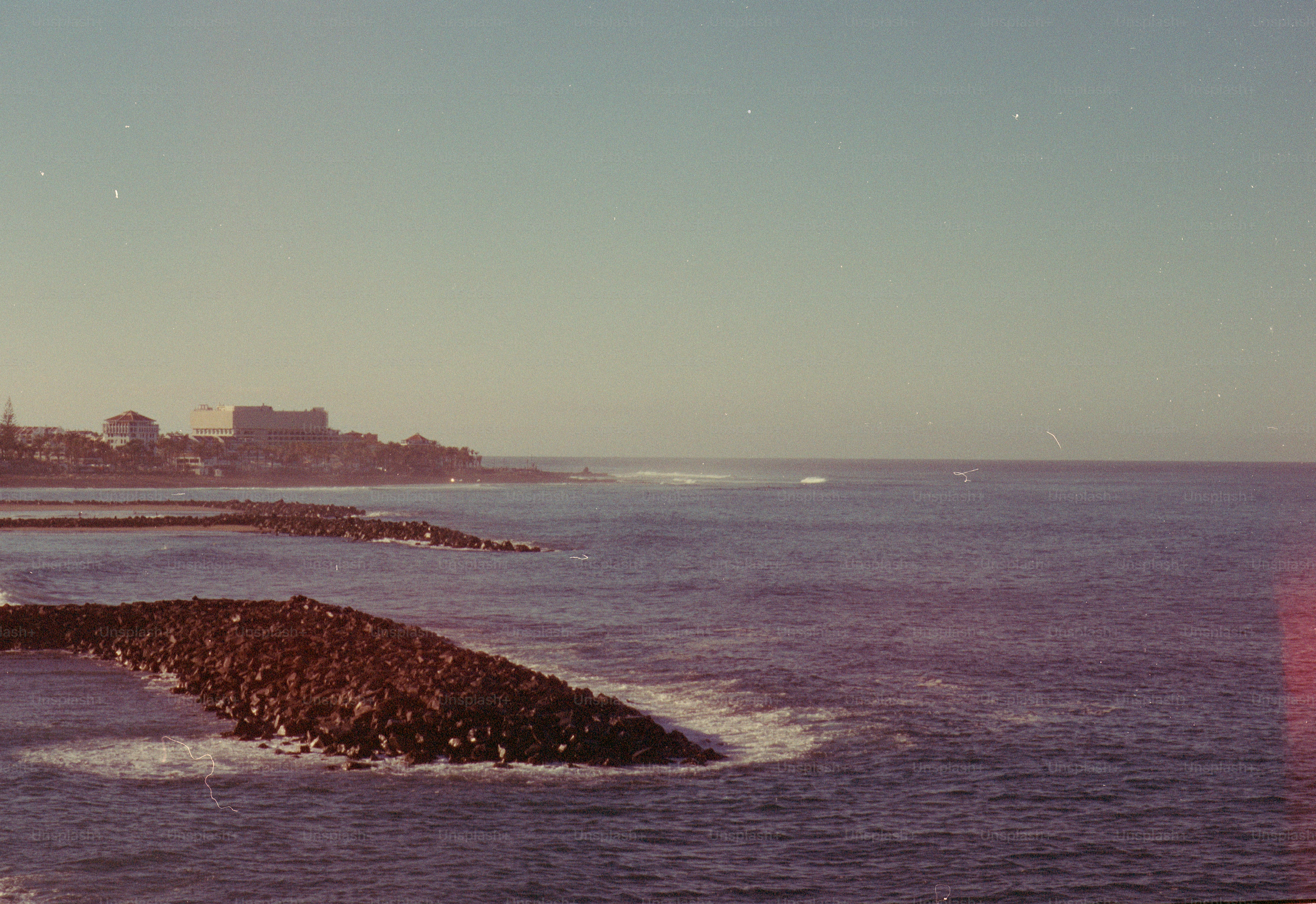Waves crash against a rocky breakwater with city skyline.