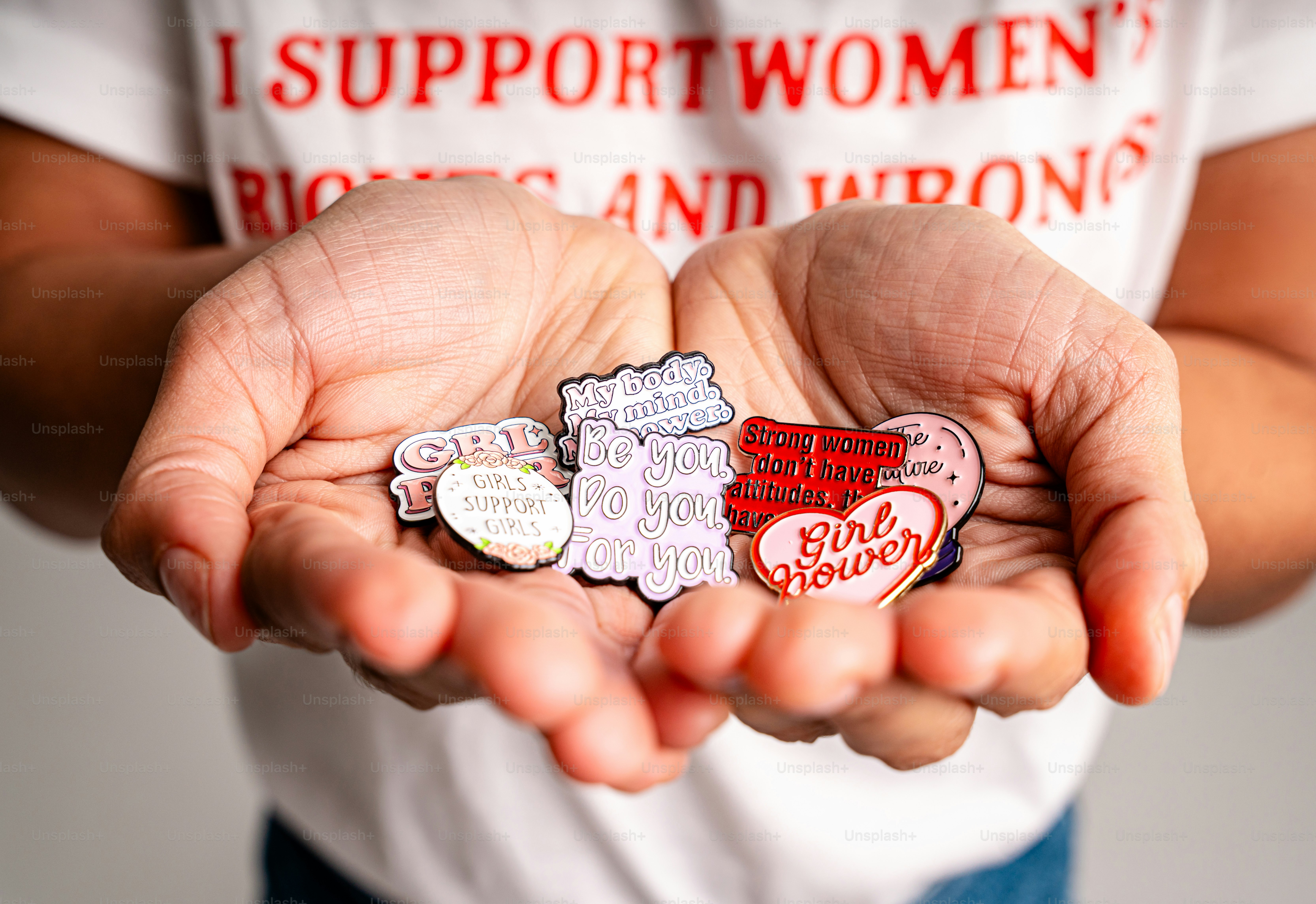 Hands holding several enamel pins with feminist slogans.