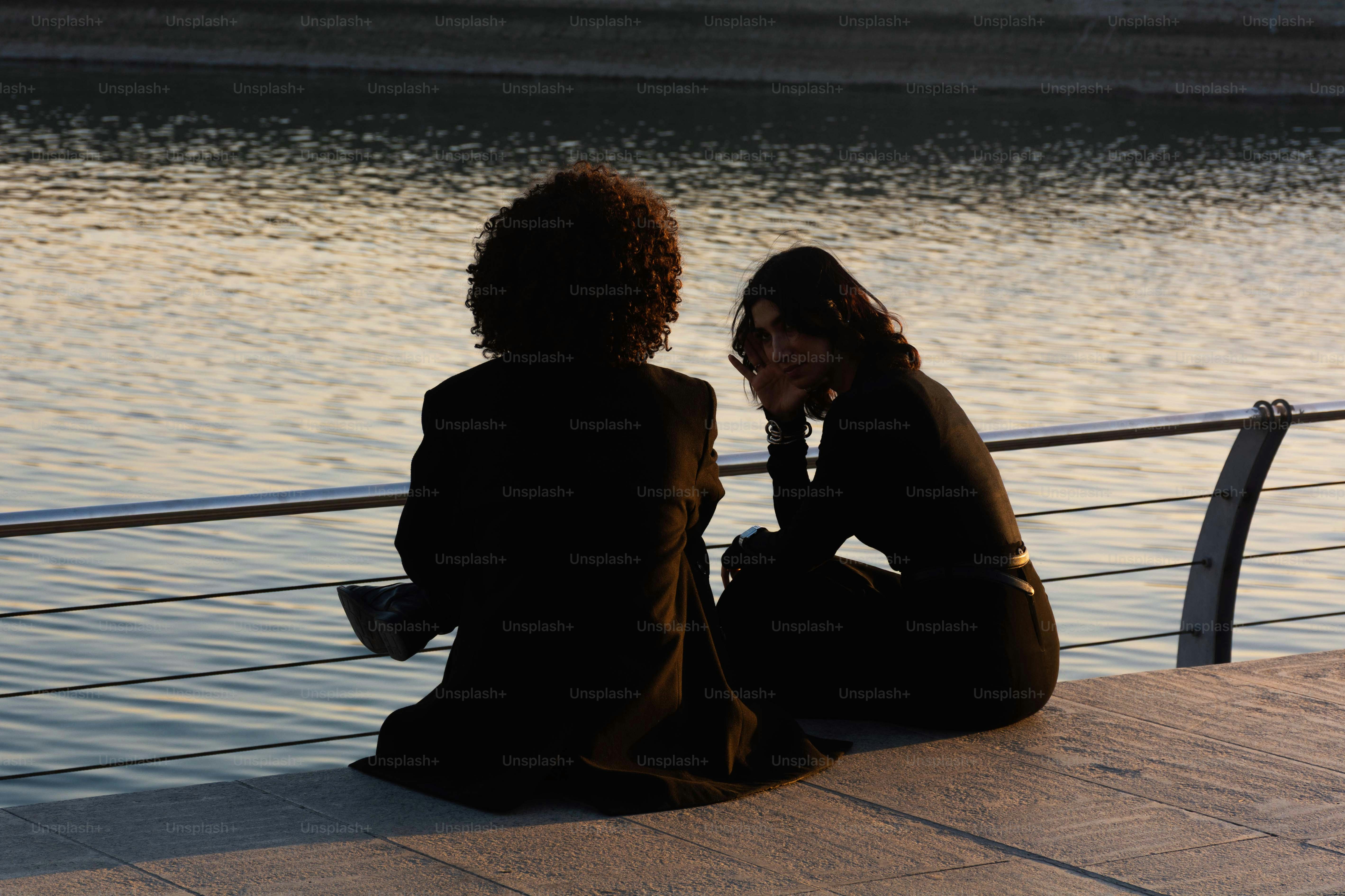 Dos personas sentadas junto al agua al atardecer.