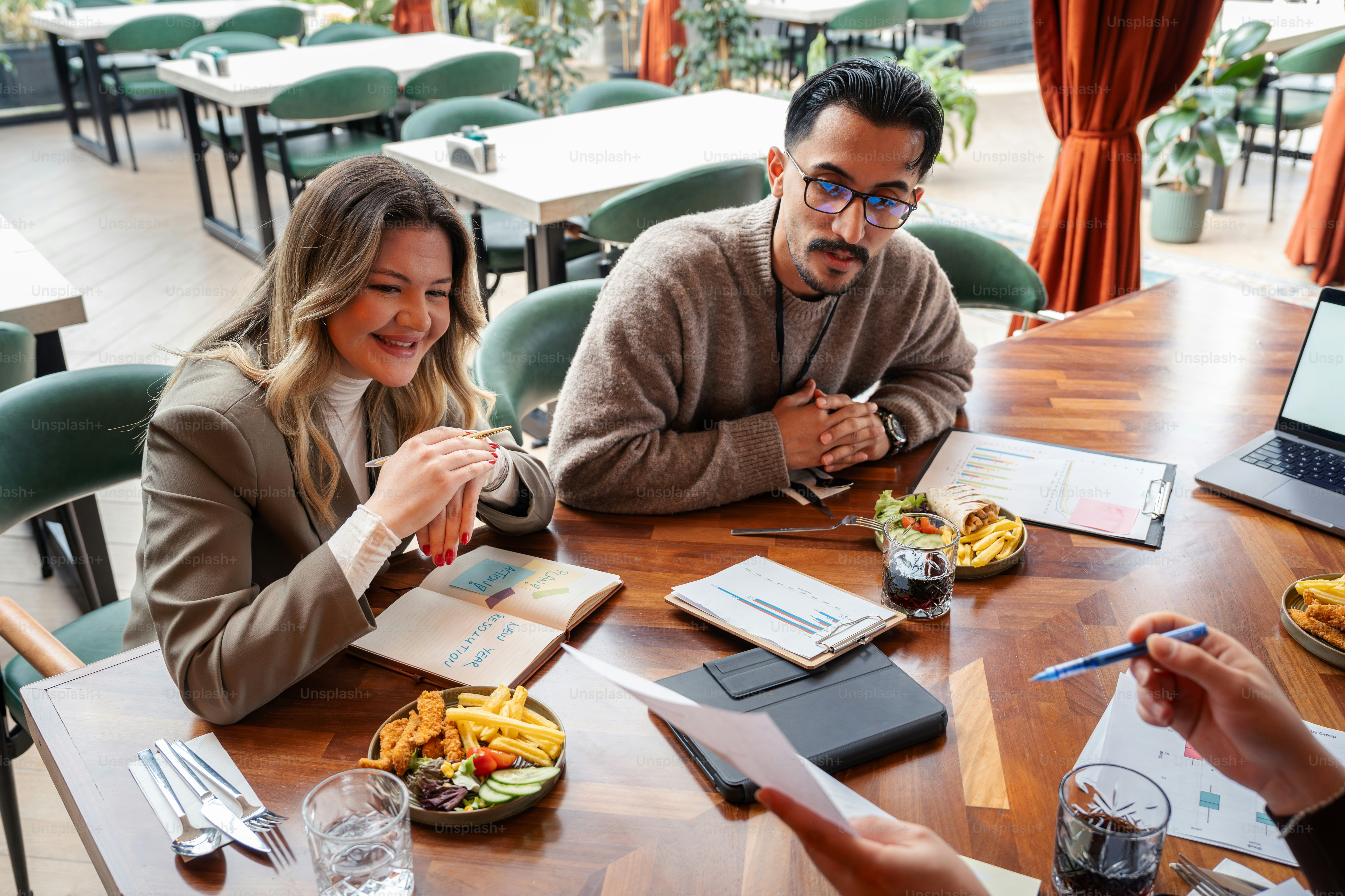 Two people sitting at a table with food and papers.