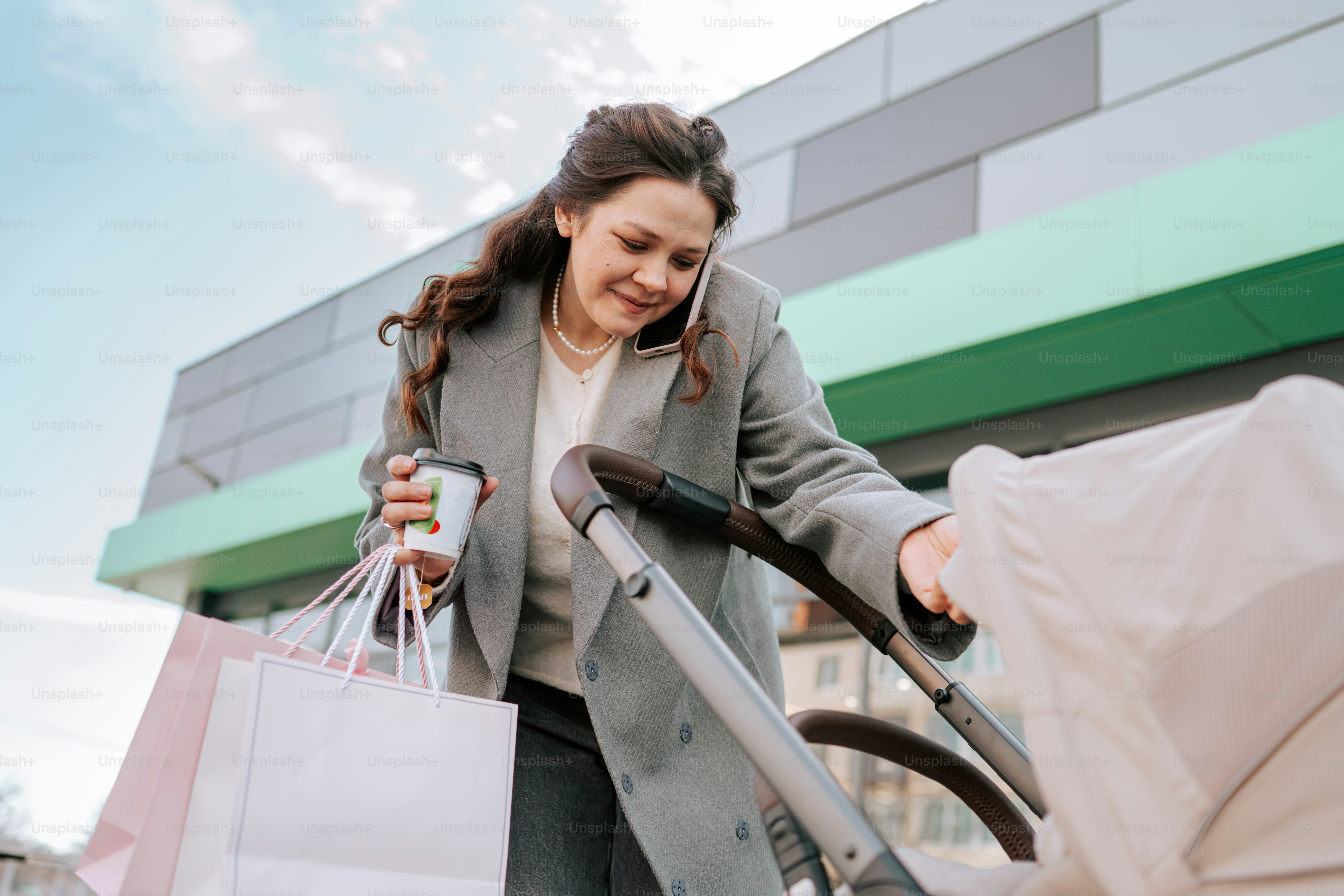 Woman with coffee, shopping bags, and stroller on phone.