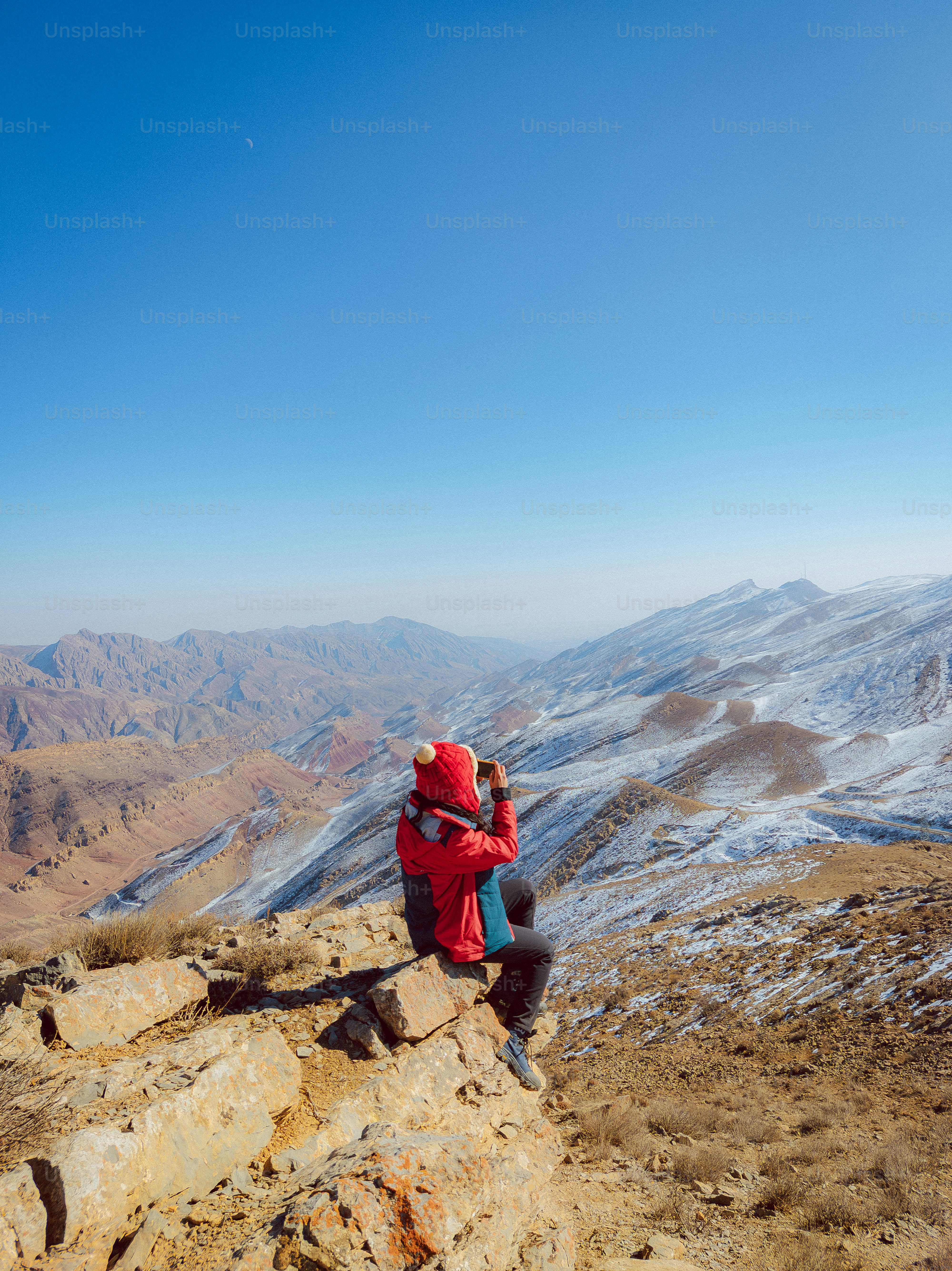 A pessoa de jaqueta vermelha senta-se em pedras com vista para montanhas nevadas.