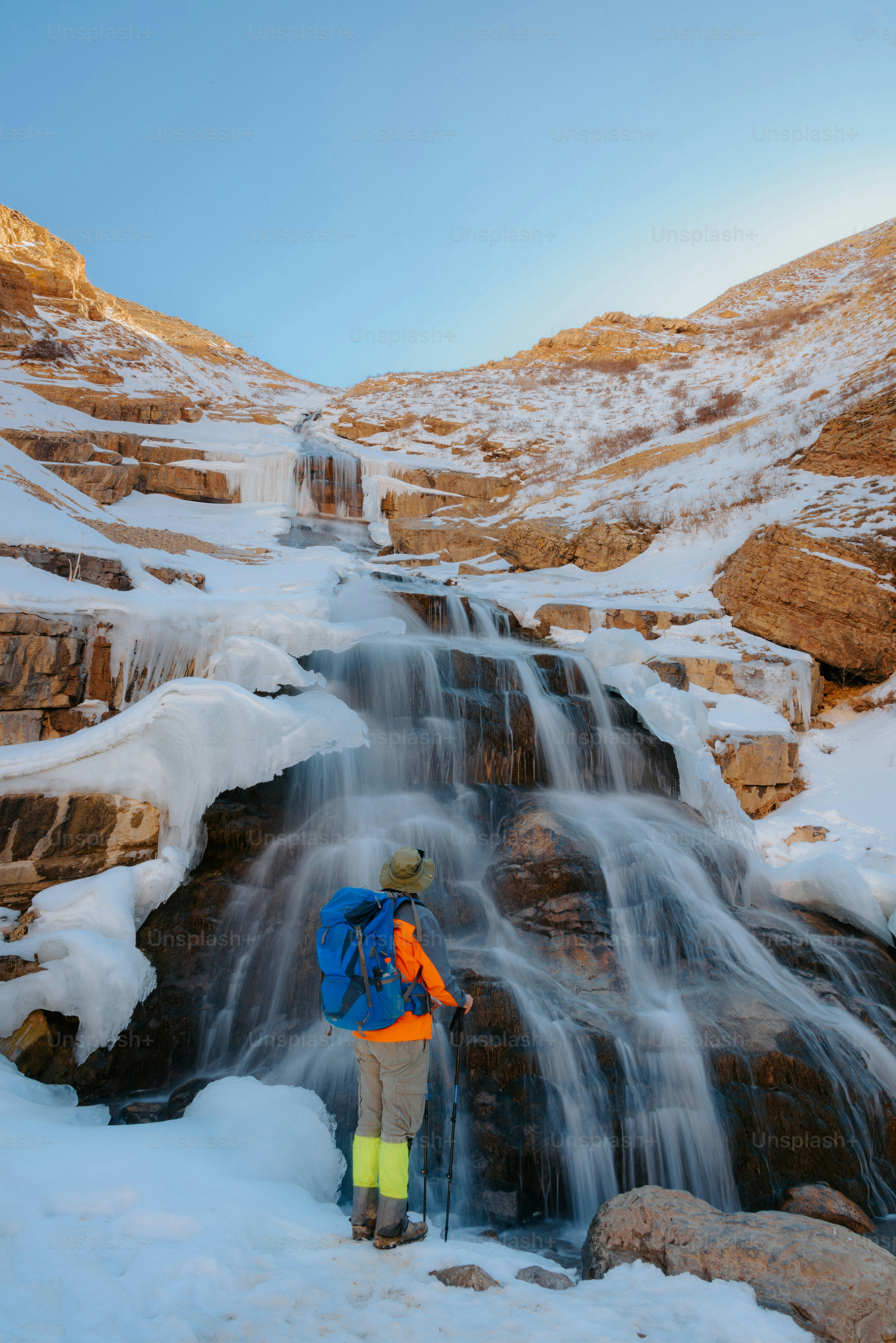Hiker observing a partially frozen waterfall in mountains.