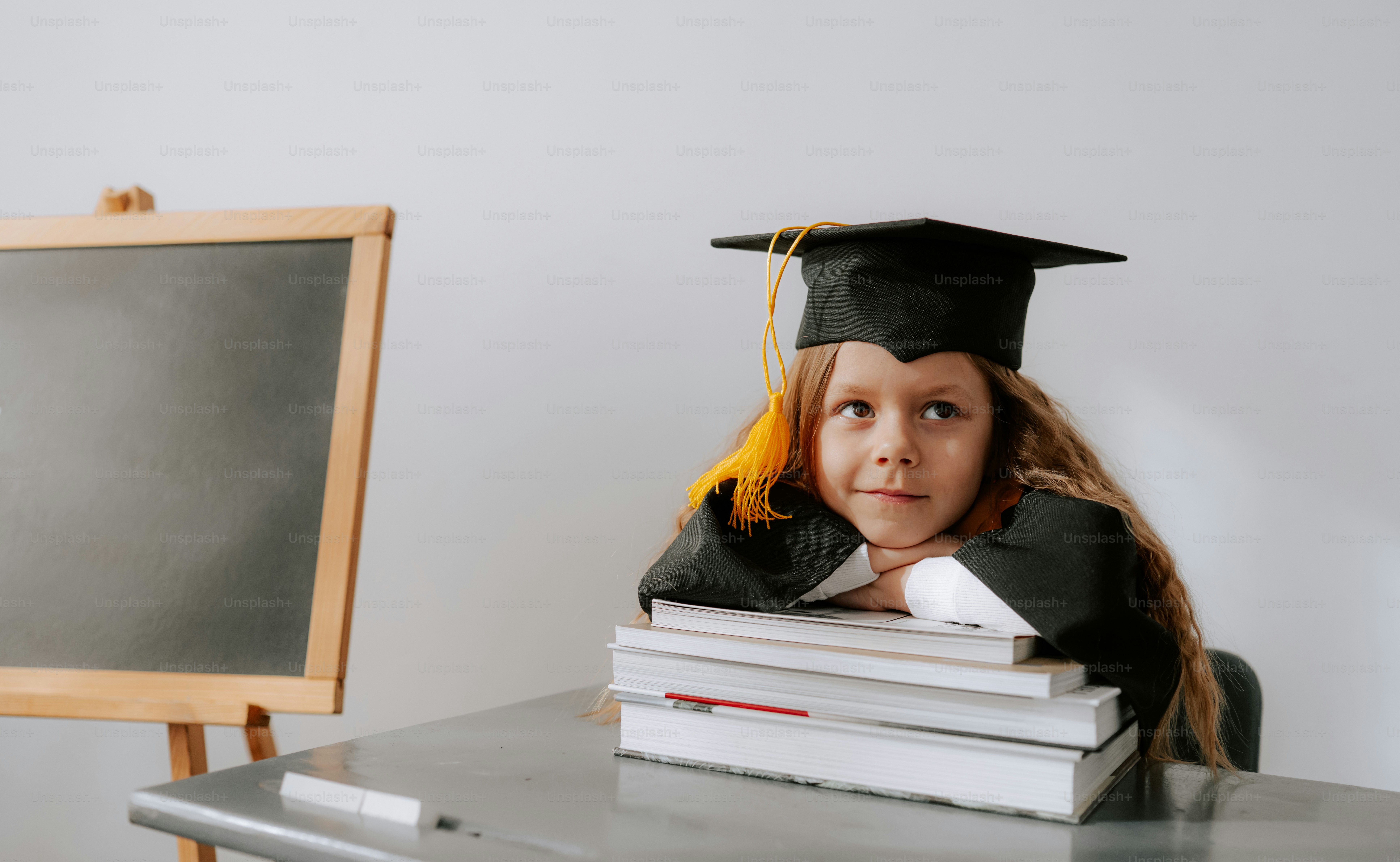 Young girl in graduation cap rests head on books.