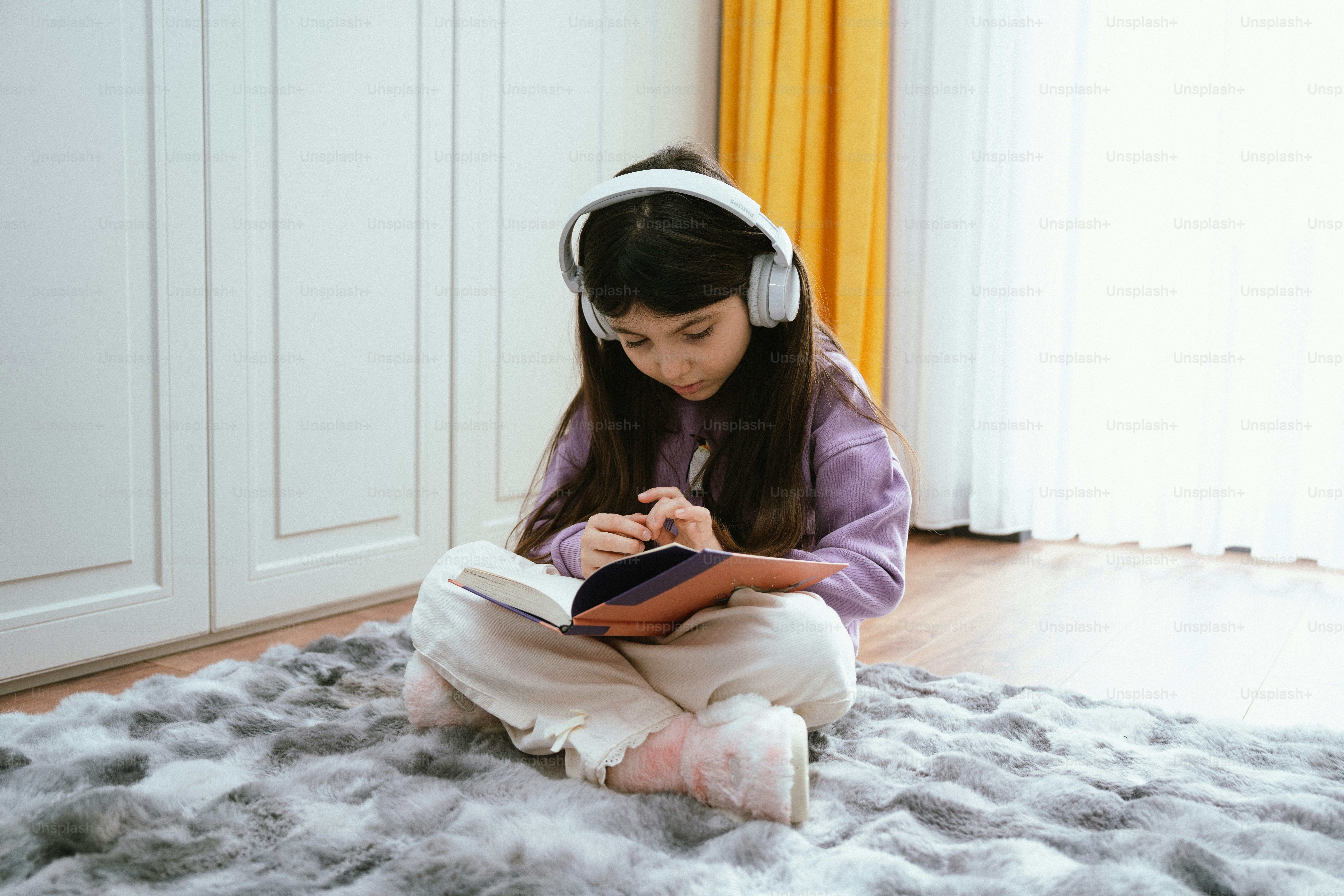 Young girl with headphones reads a book on rug