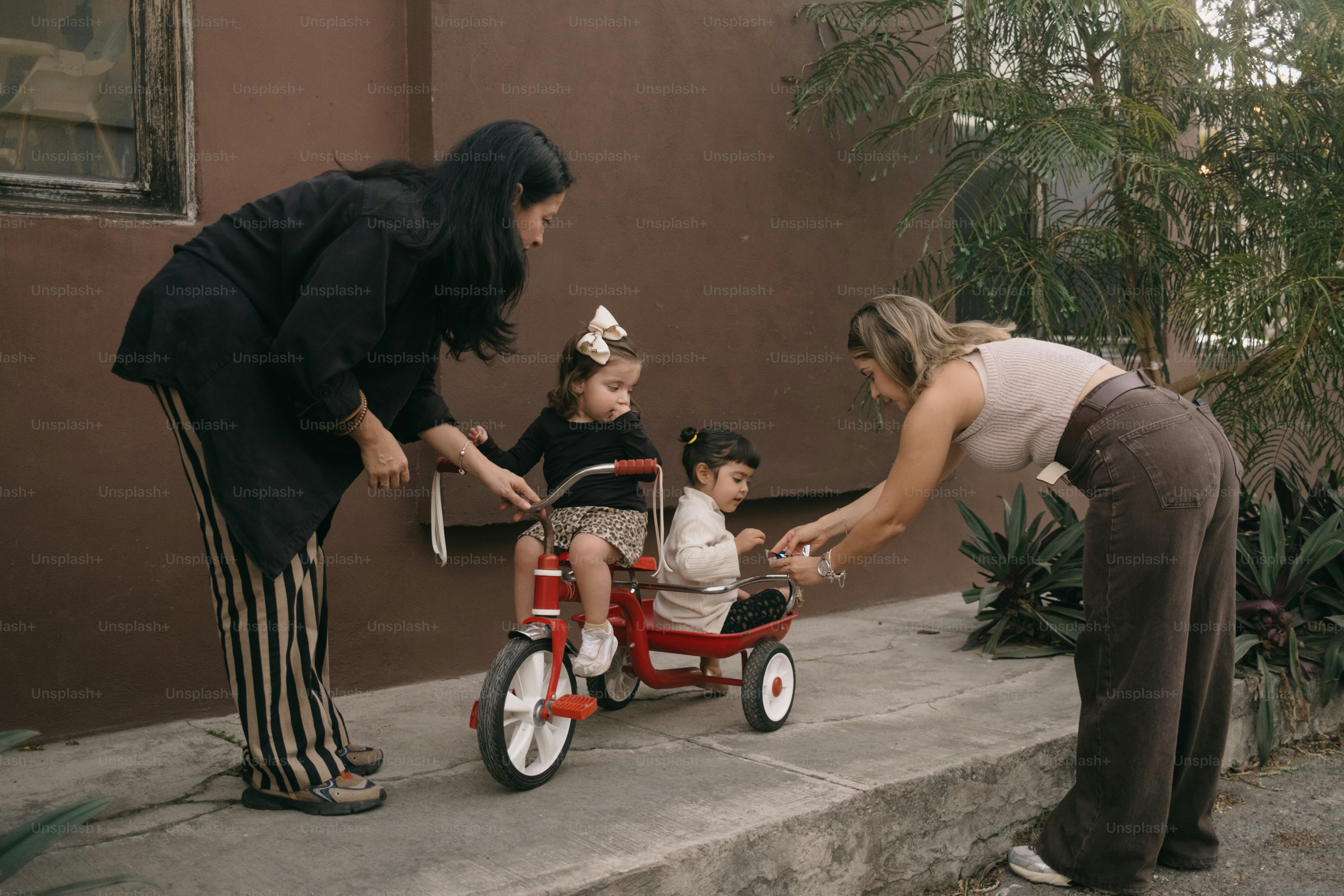 Deux femmes aident deux jeunes enfants sur un tricycle.