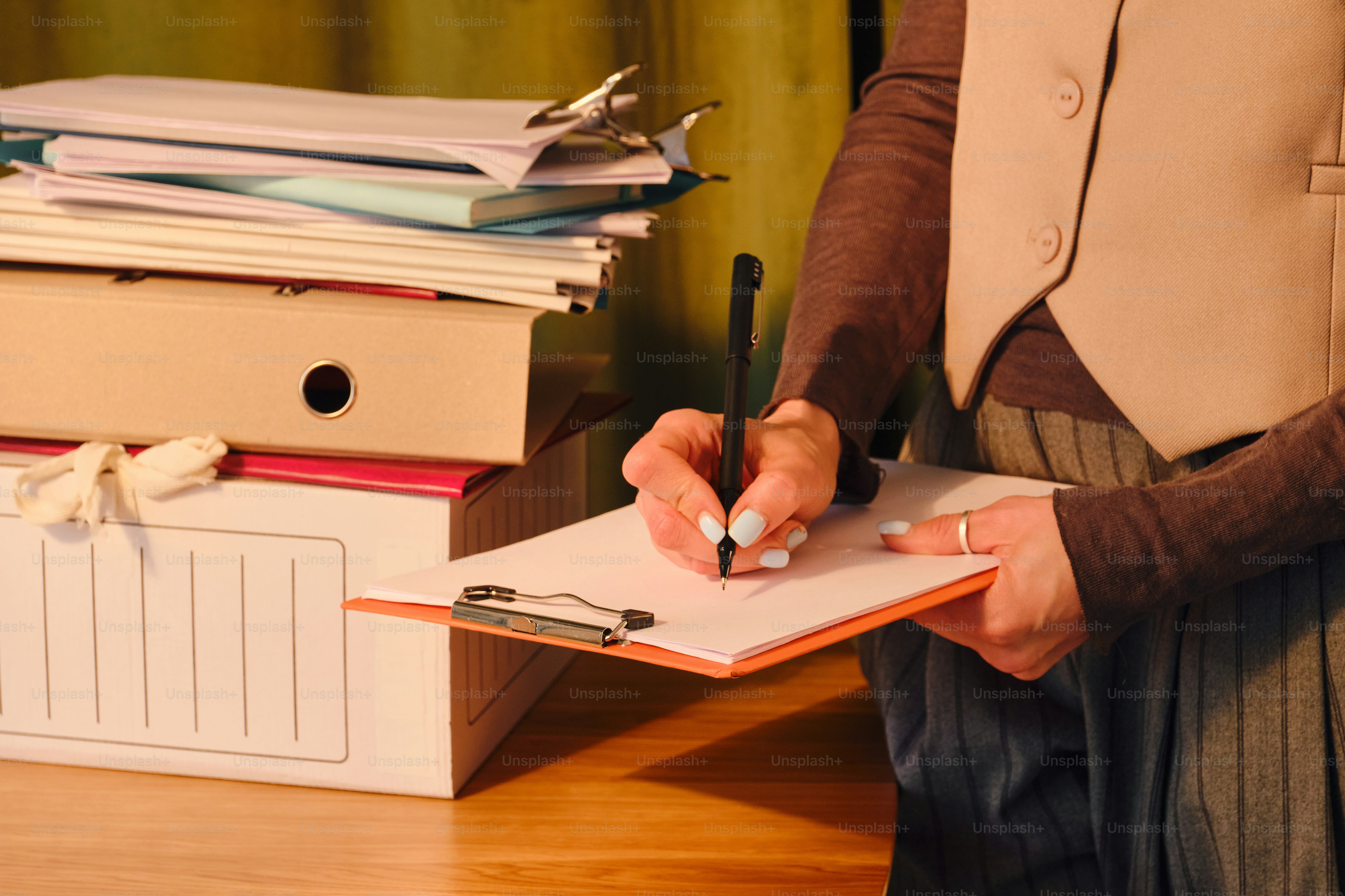 Person writing on clipboard near stack of documents.
