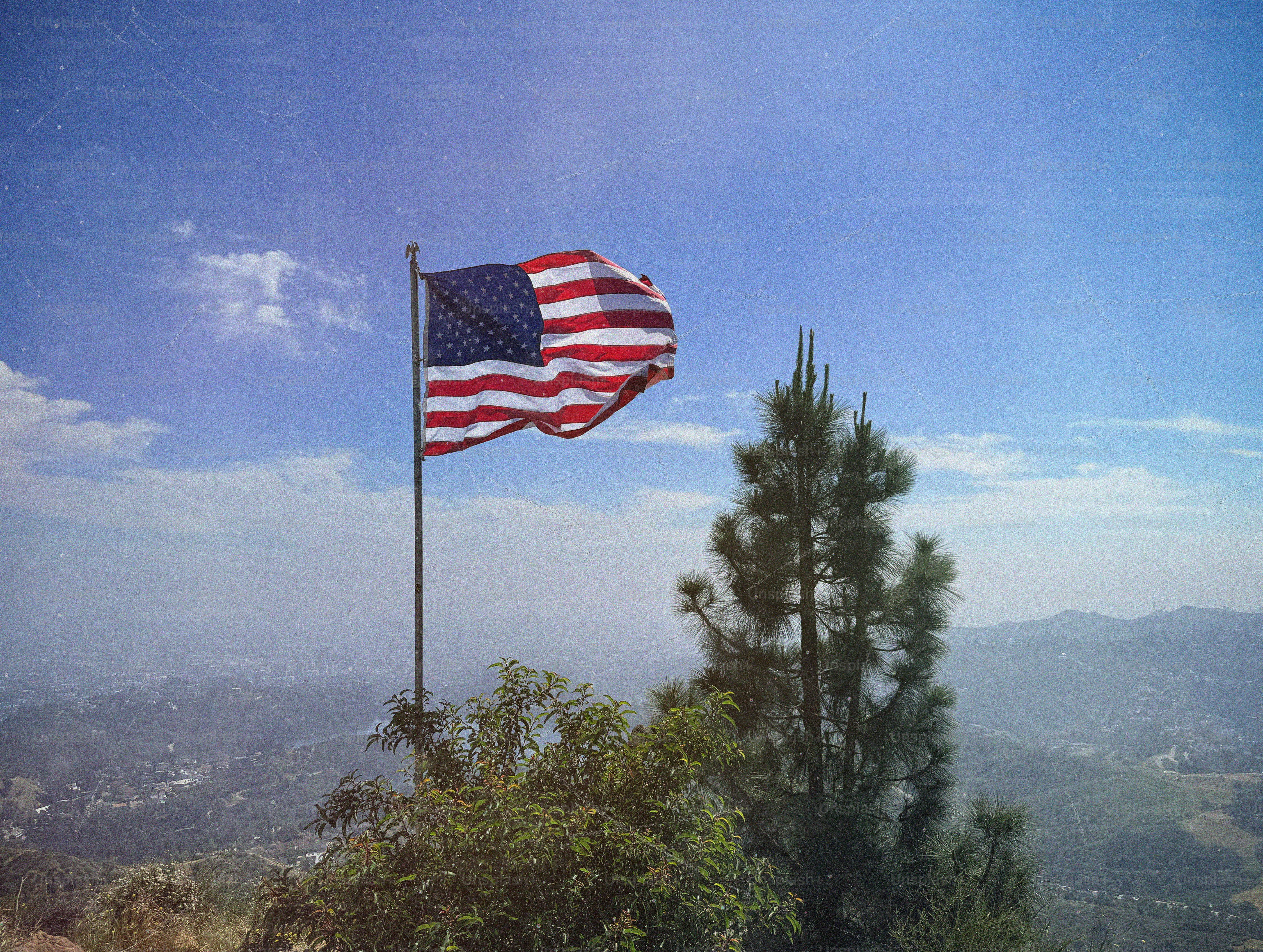 American flag flying on a pole with trees and hills.