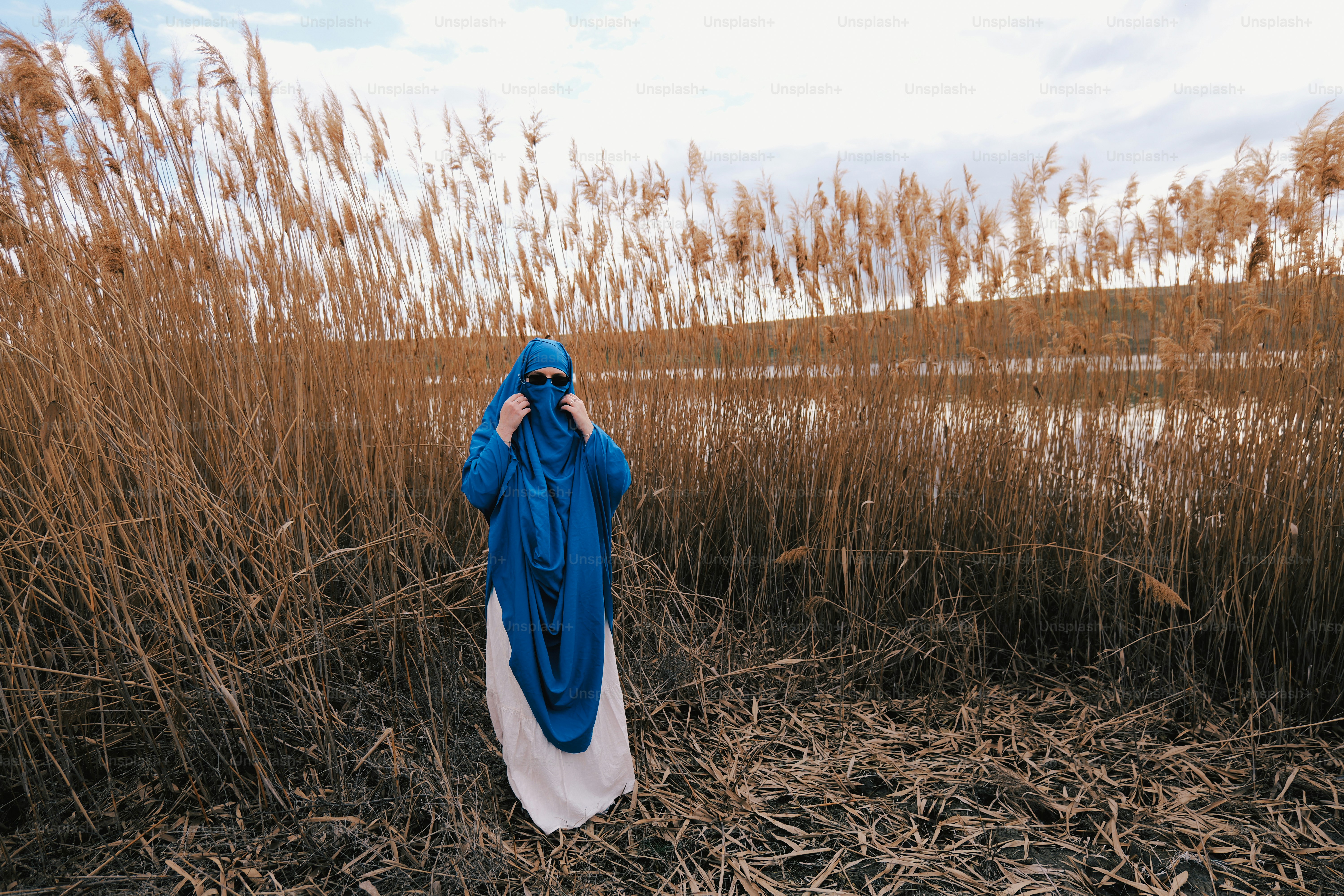 Wanita berjilbab biru berdiri di alang-alang kering di tepi air