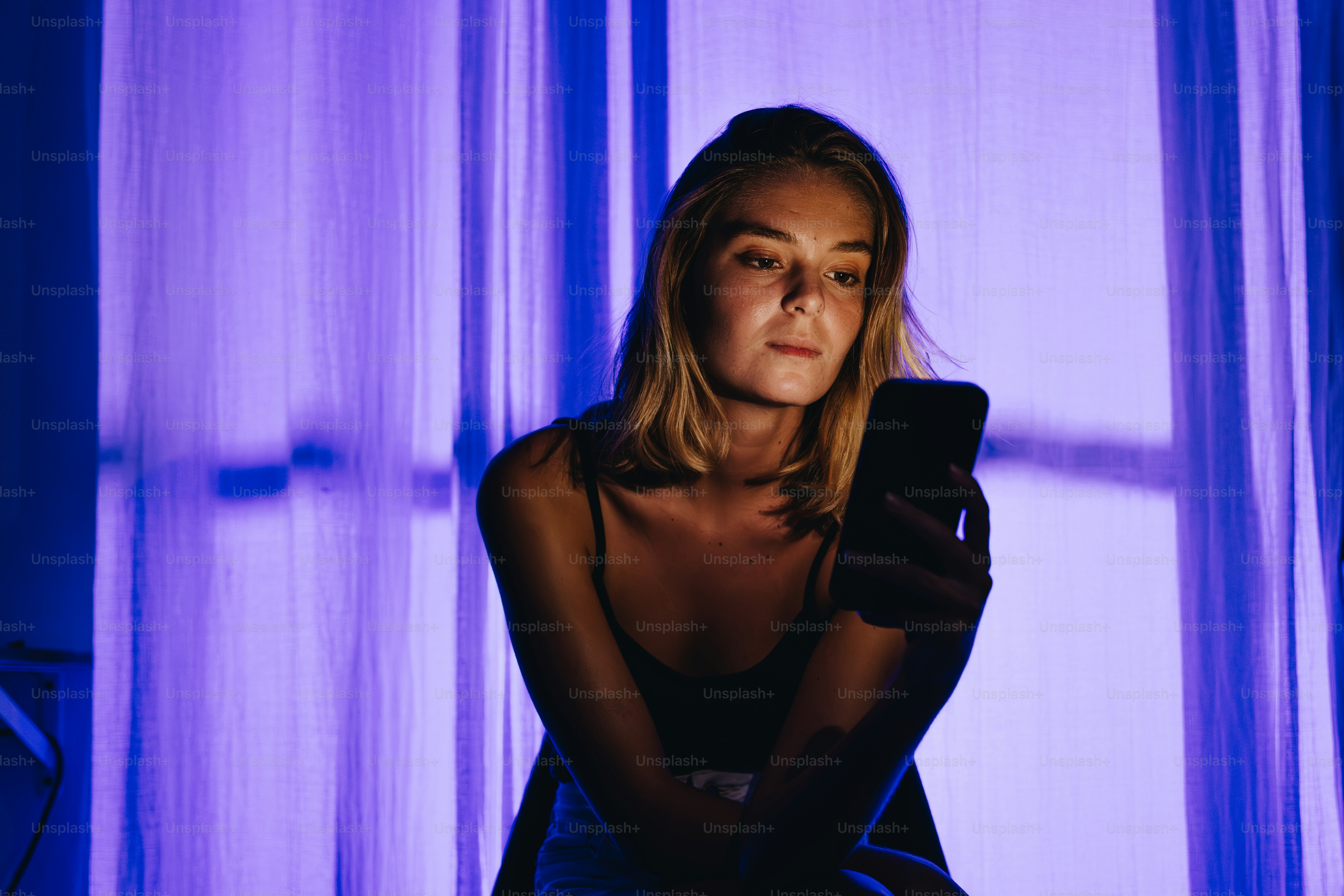 Mujer iluminada por la pantalla del móvil contra cortinas moradas