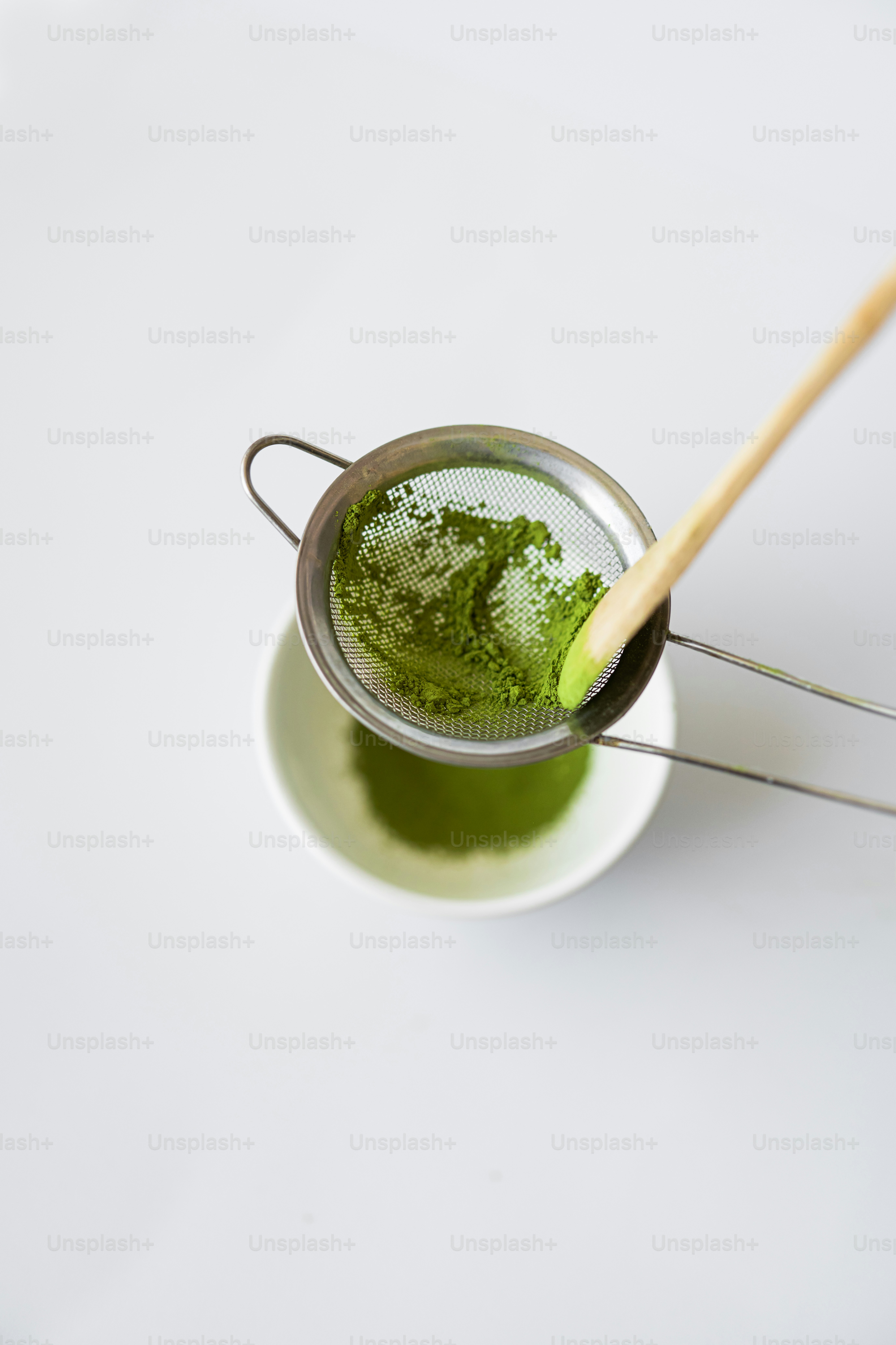 Bubuk matcha hijau diayak ke dalam mangkuk putih.