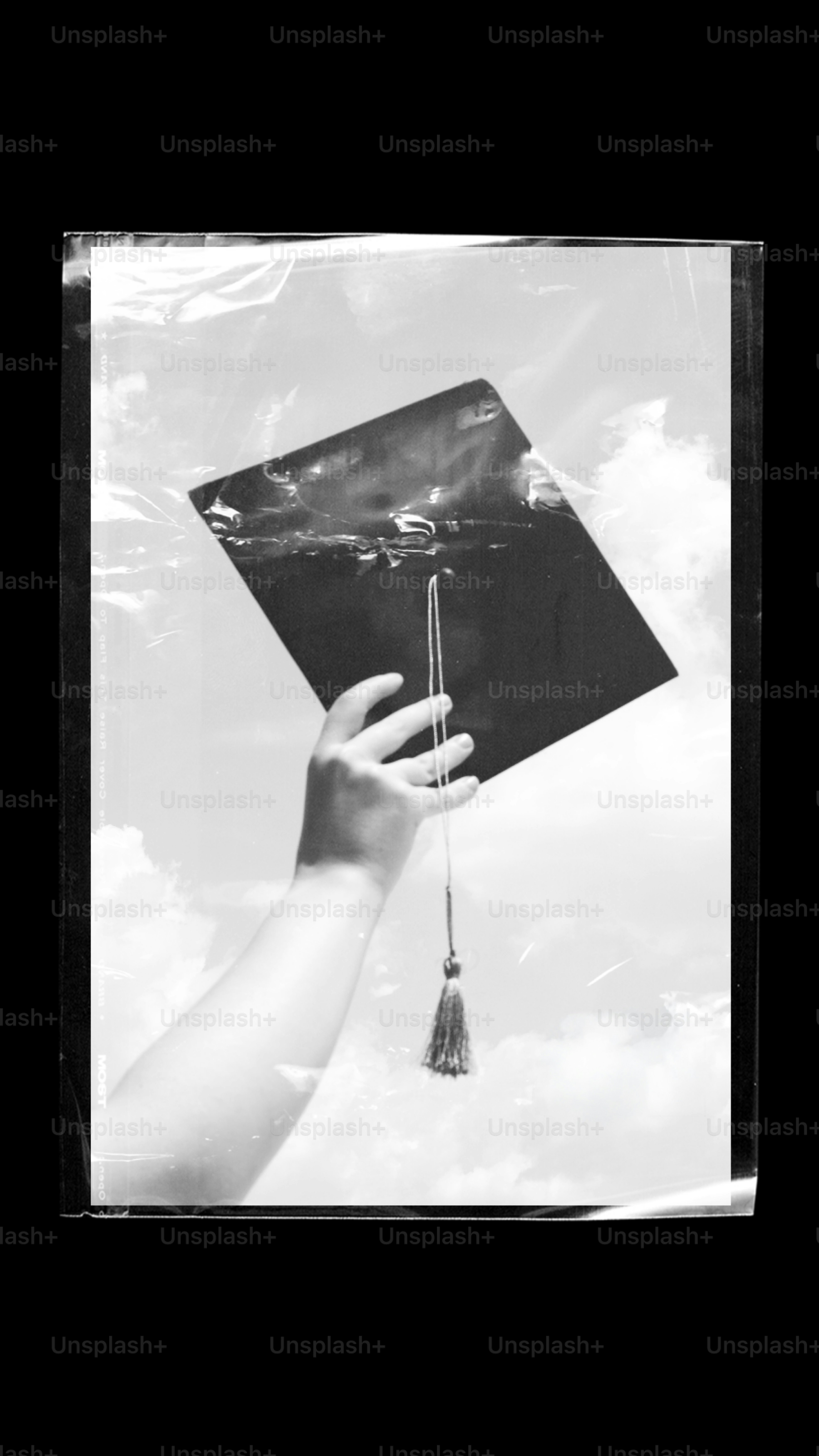 Hand holding graduation cap against cloudy sky.