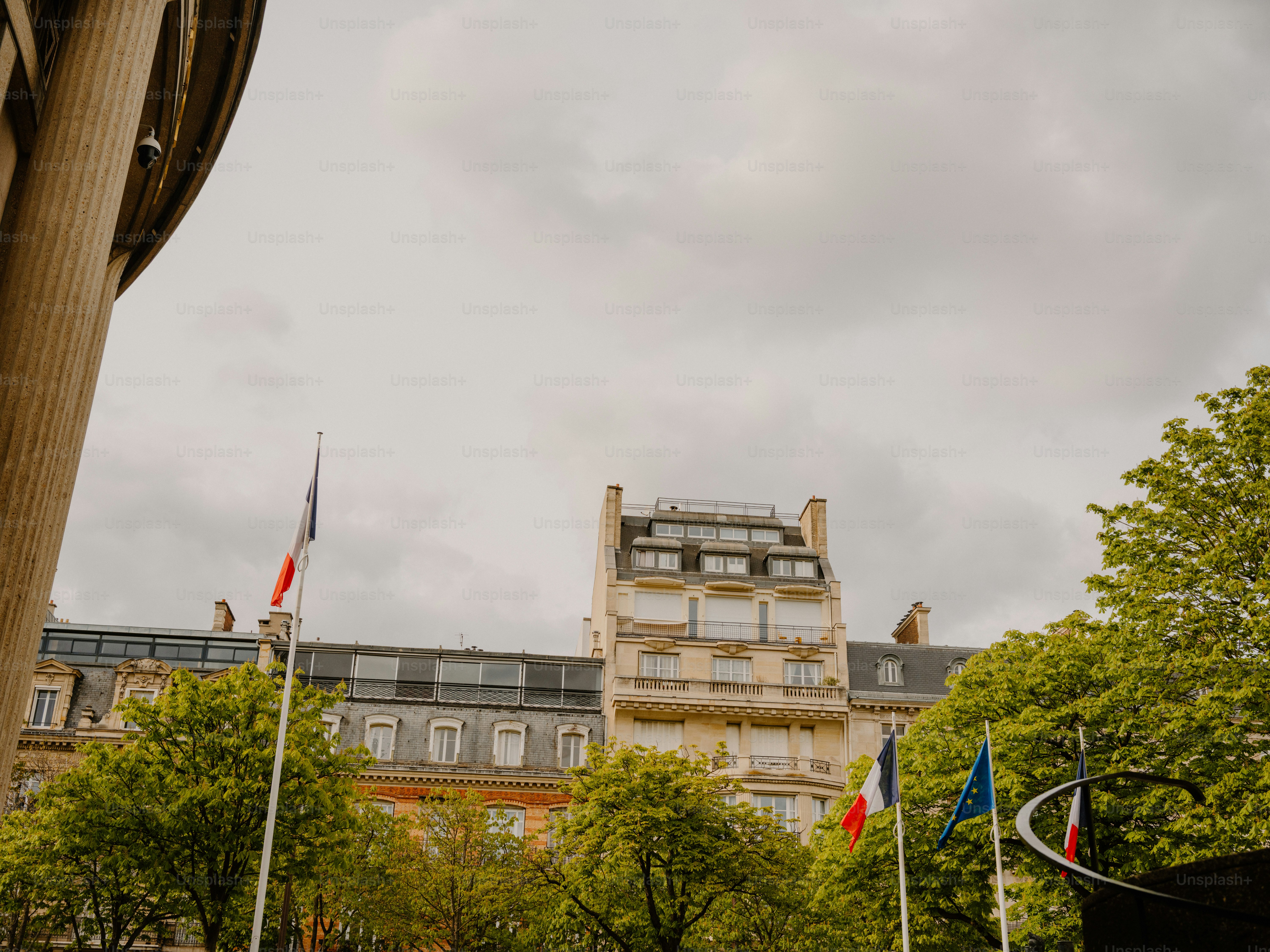 French flags fly before a building with trees.