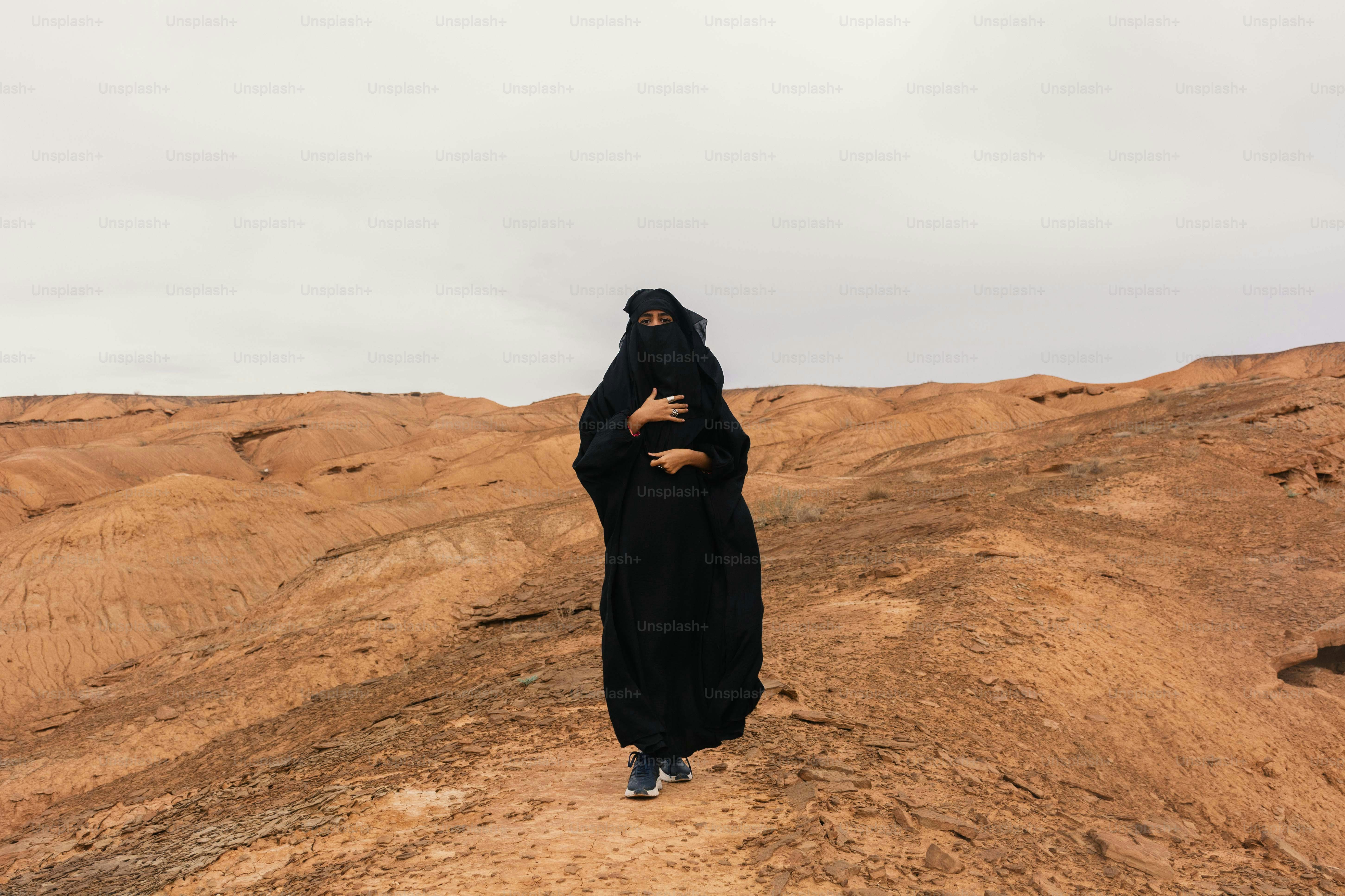 Wanita berbaju abaya hitam berdiri di bukit pasir gurun