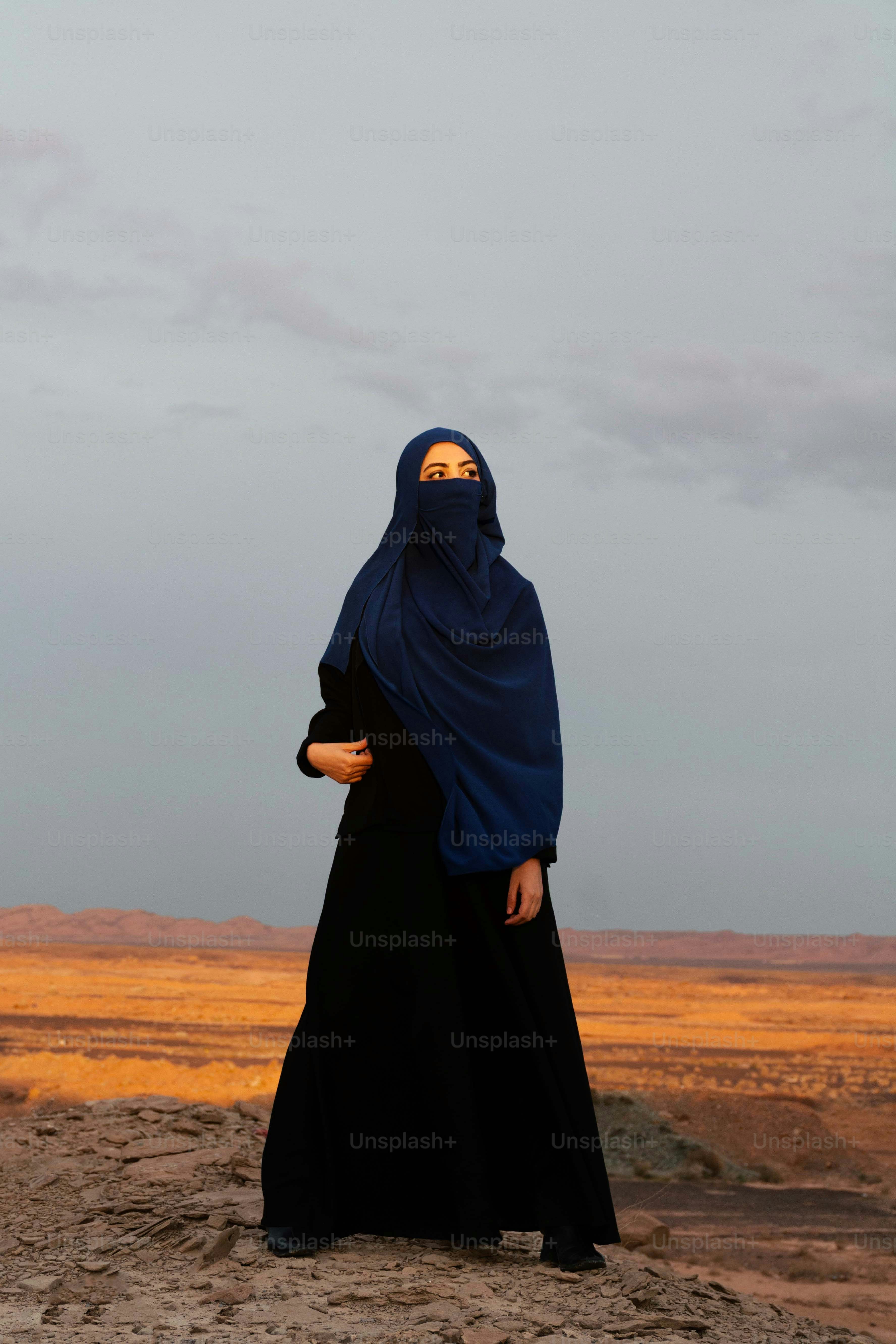 Wanita dalam niqab dan abaya berdiri di lanskap gurun berbatu.