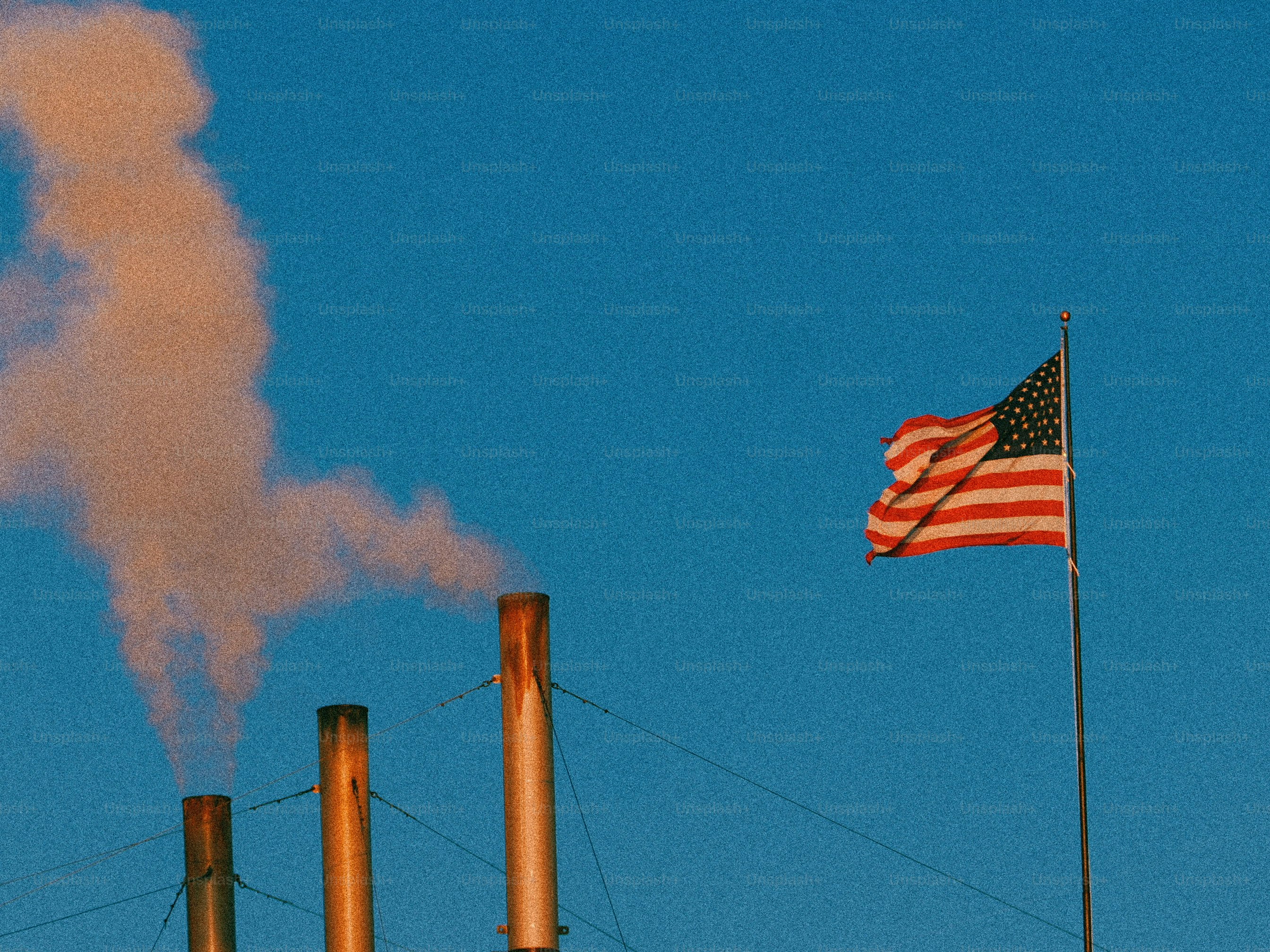 American flag waves near smoking industrial smokestacks.