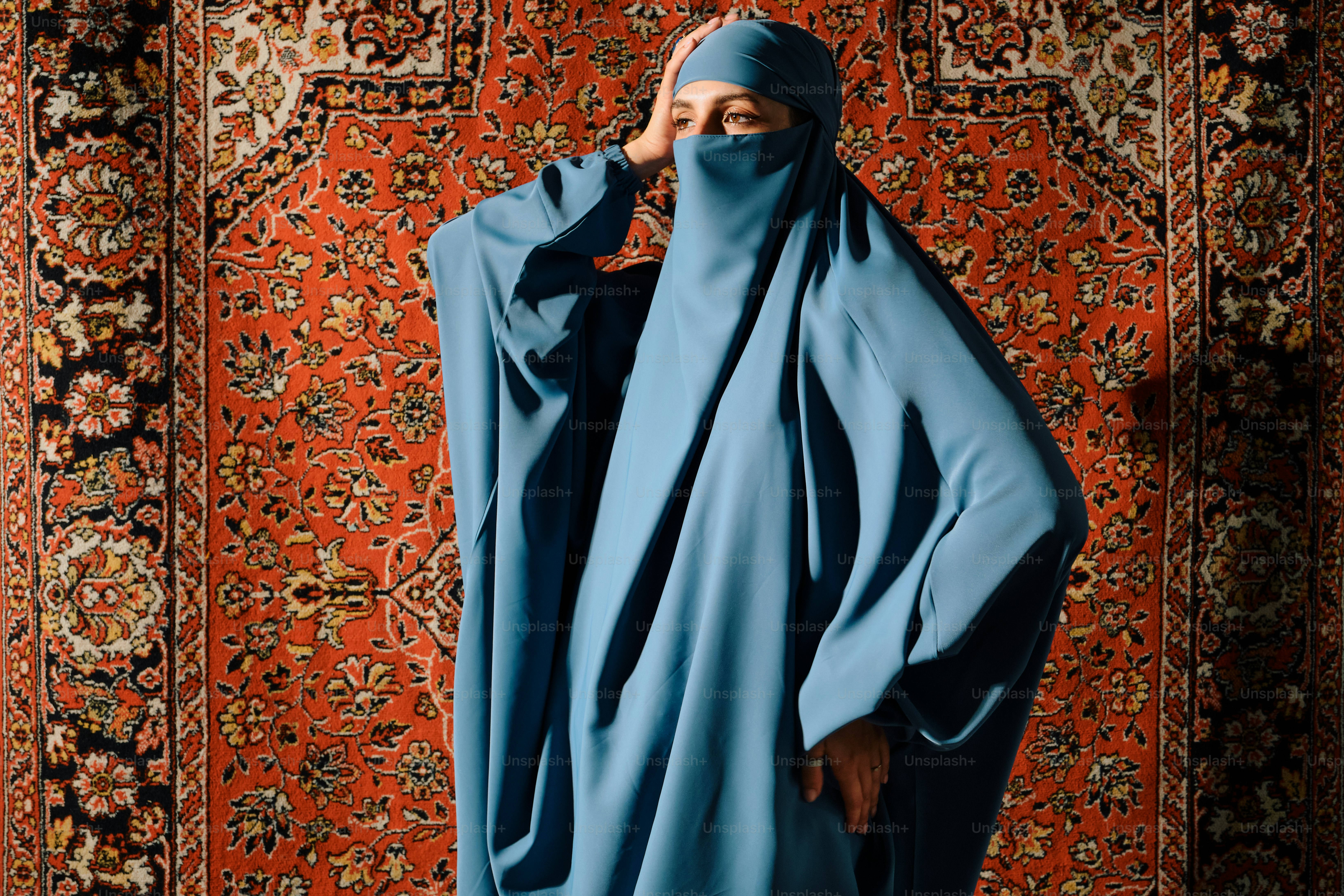 Wanita berjilbab biru dan niqab berdiri di atas karpet.