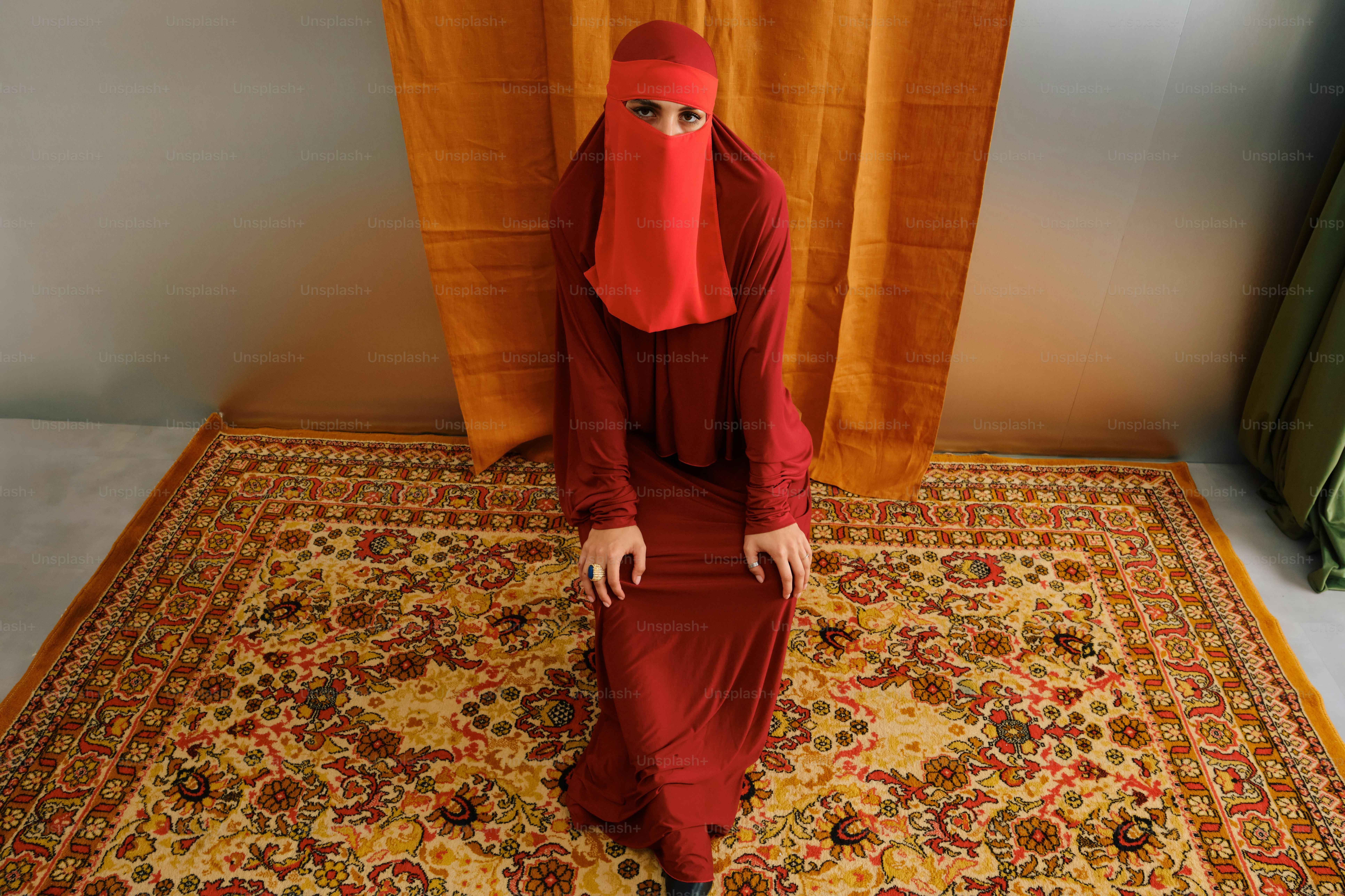 Wanita dengan niqab merah dan berpakaian di atas karpet berhias.