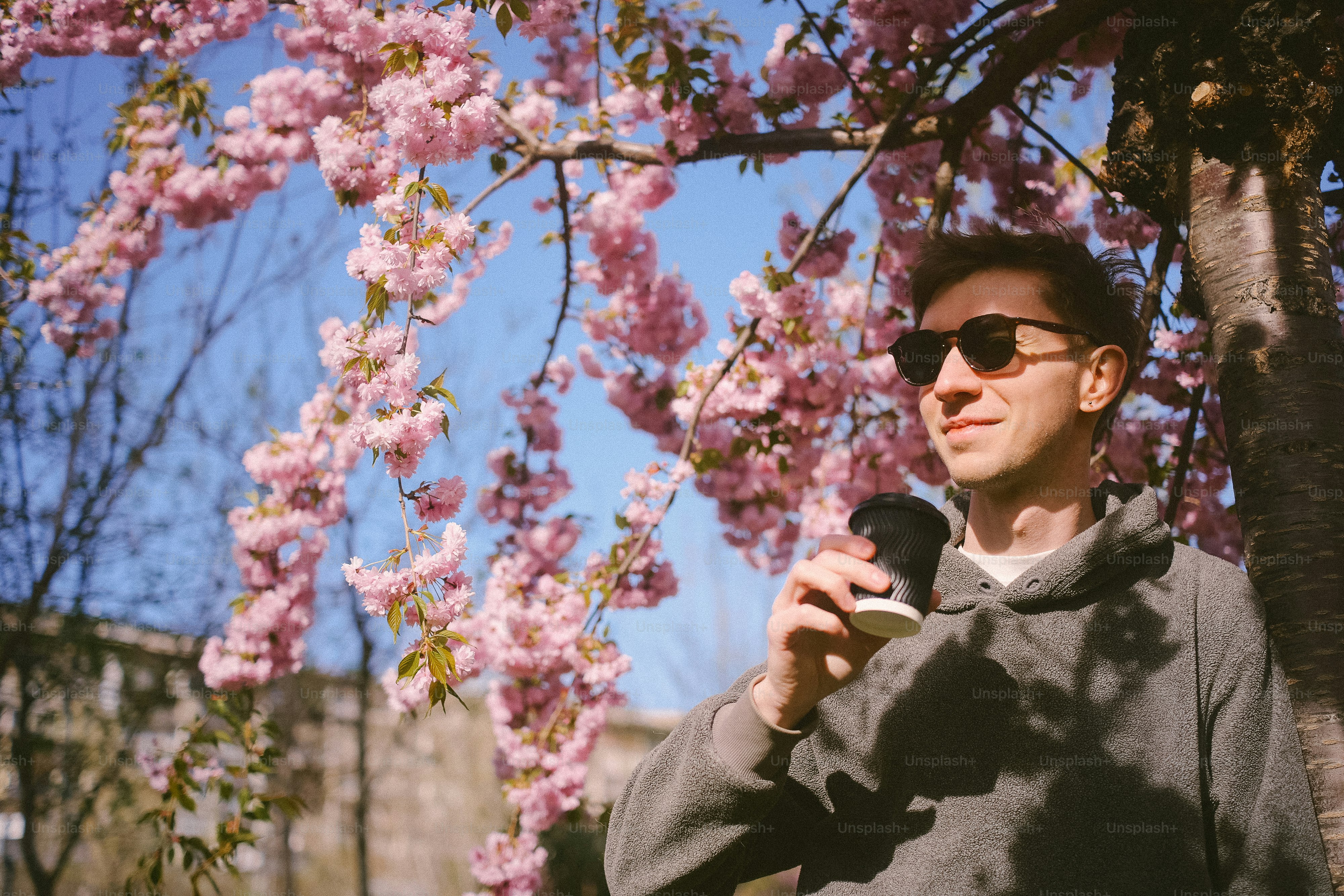 Man in sunglasses drinks coffee under cherry blossoms.