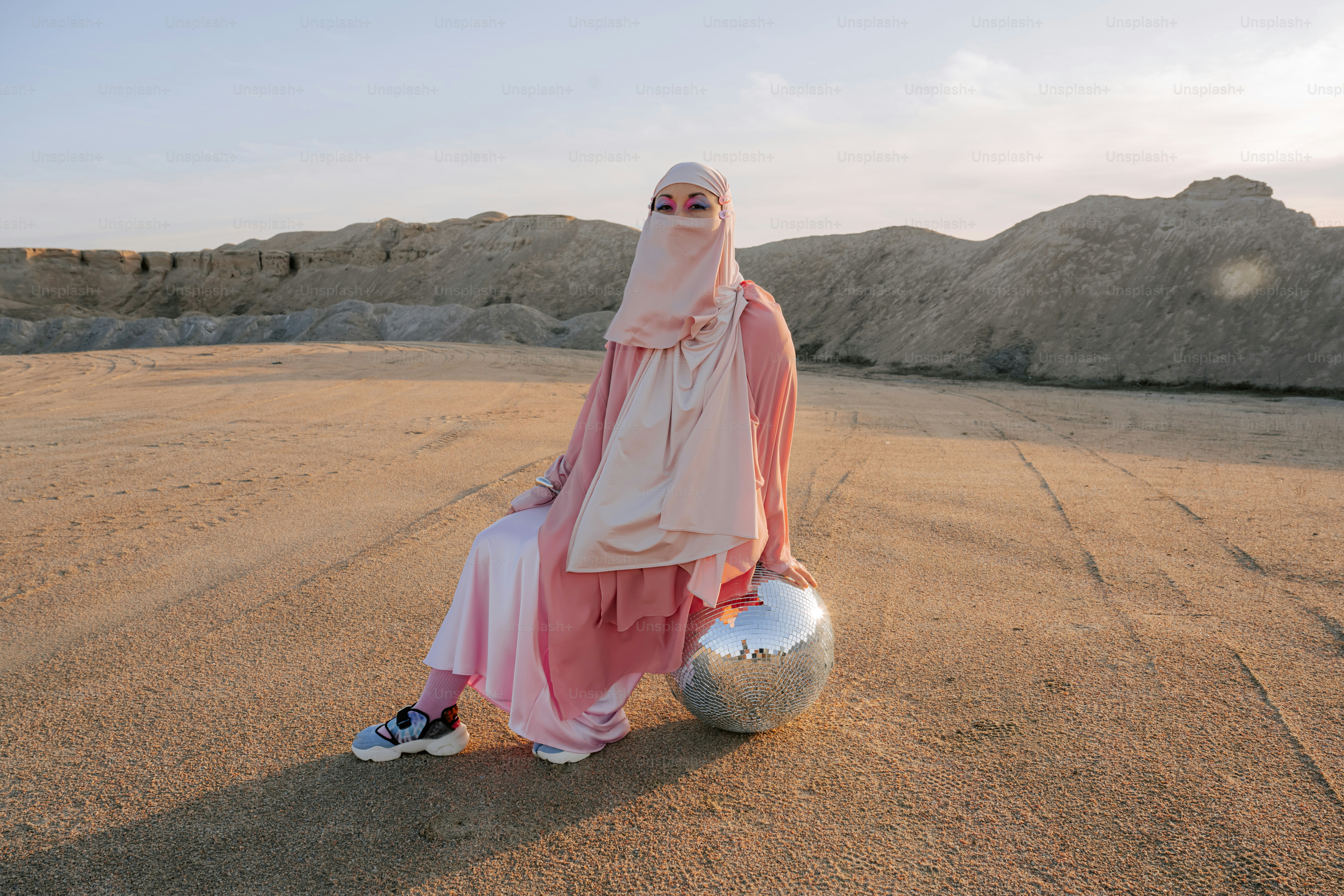 Woman in pink hijab sits with disco ball in desert.