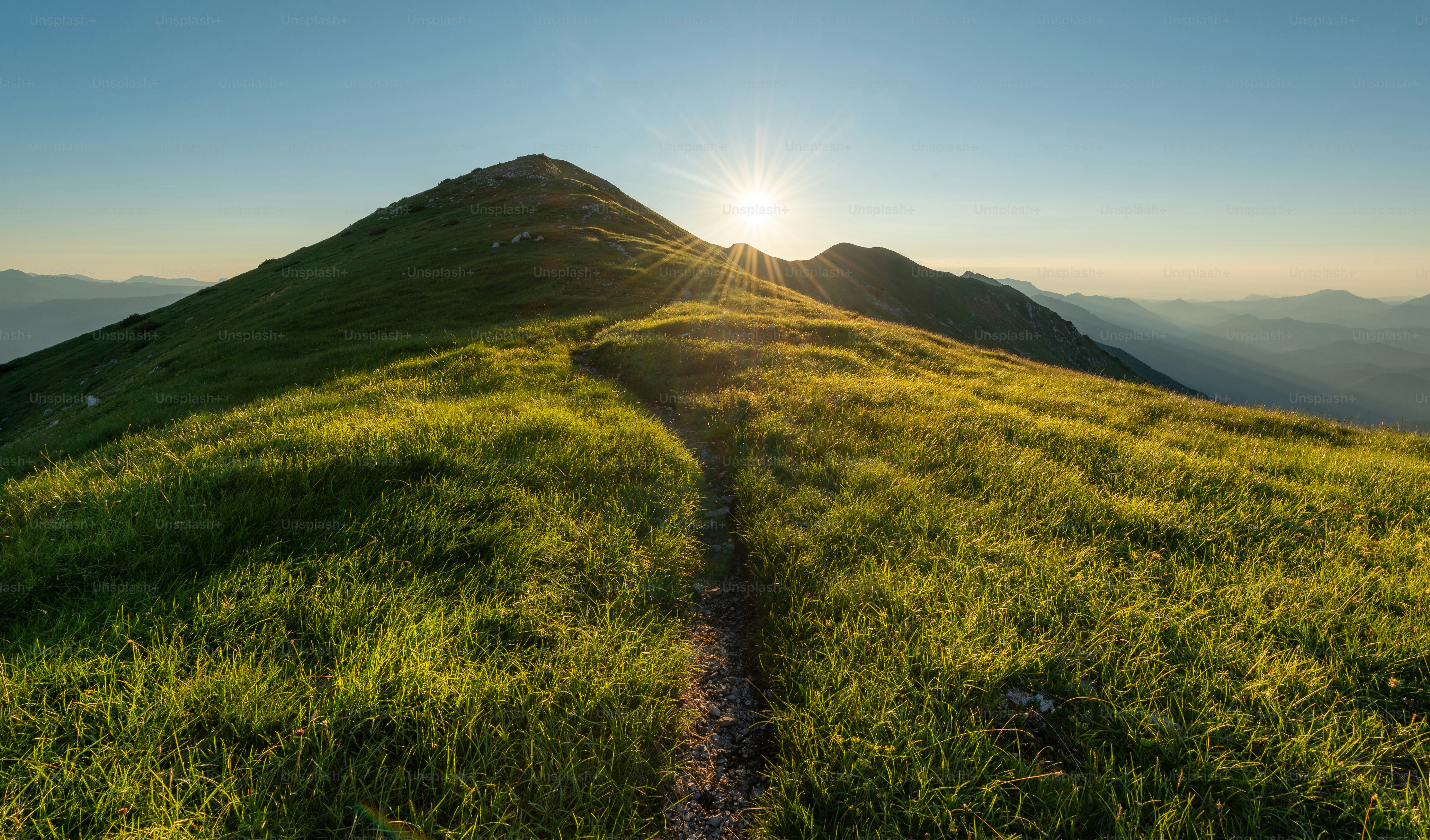 A winding path leads up a grassy mountain at sunrise.