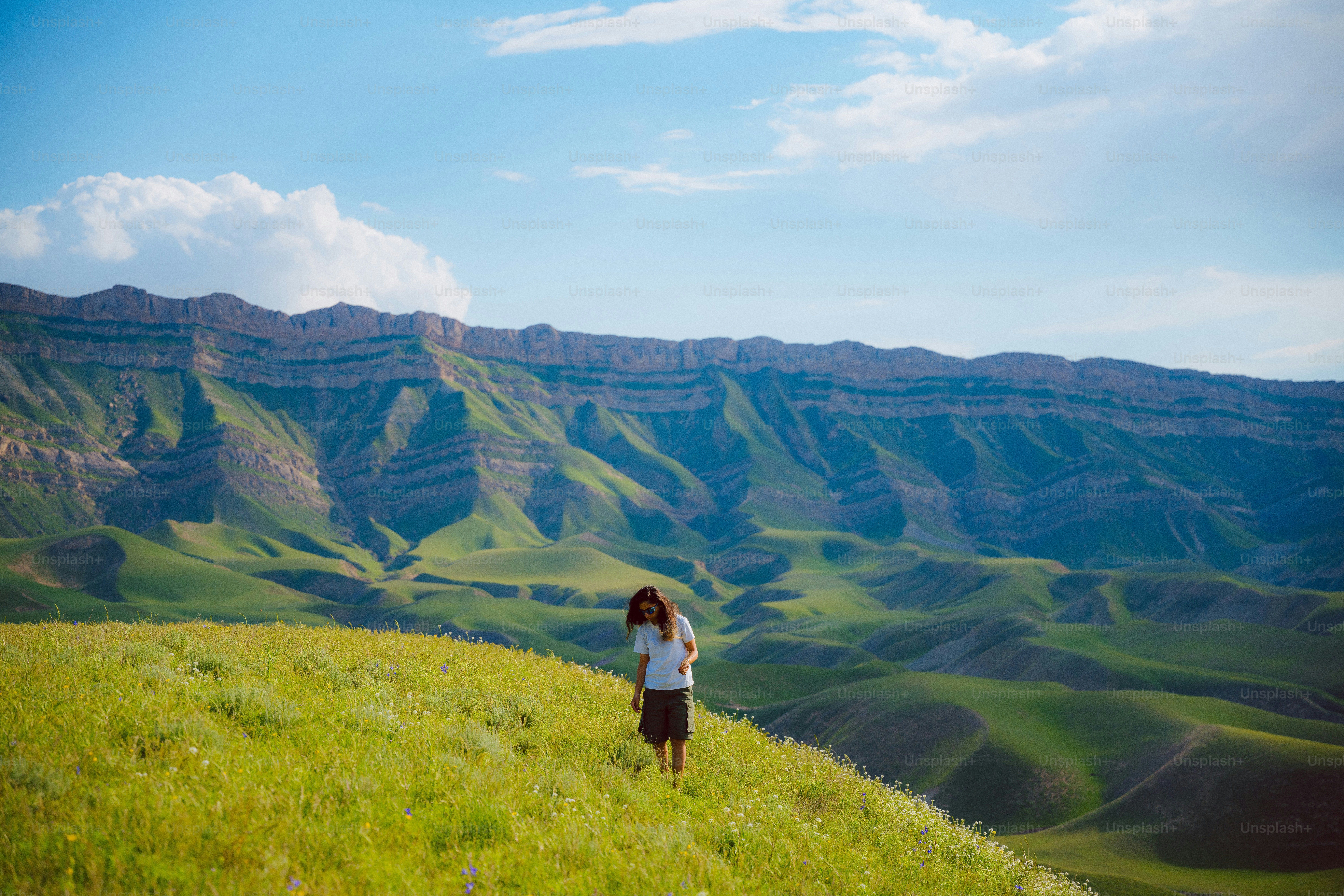 Uma pessoa caminha por uma colina gramada com montanhas atrás.