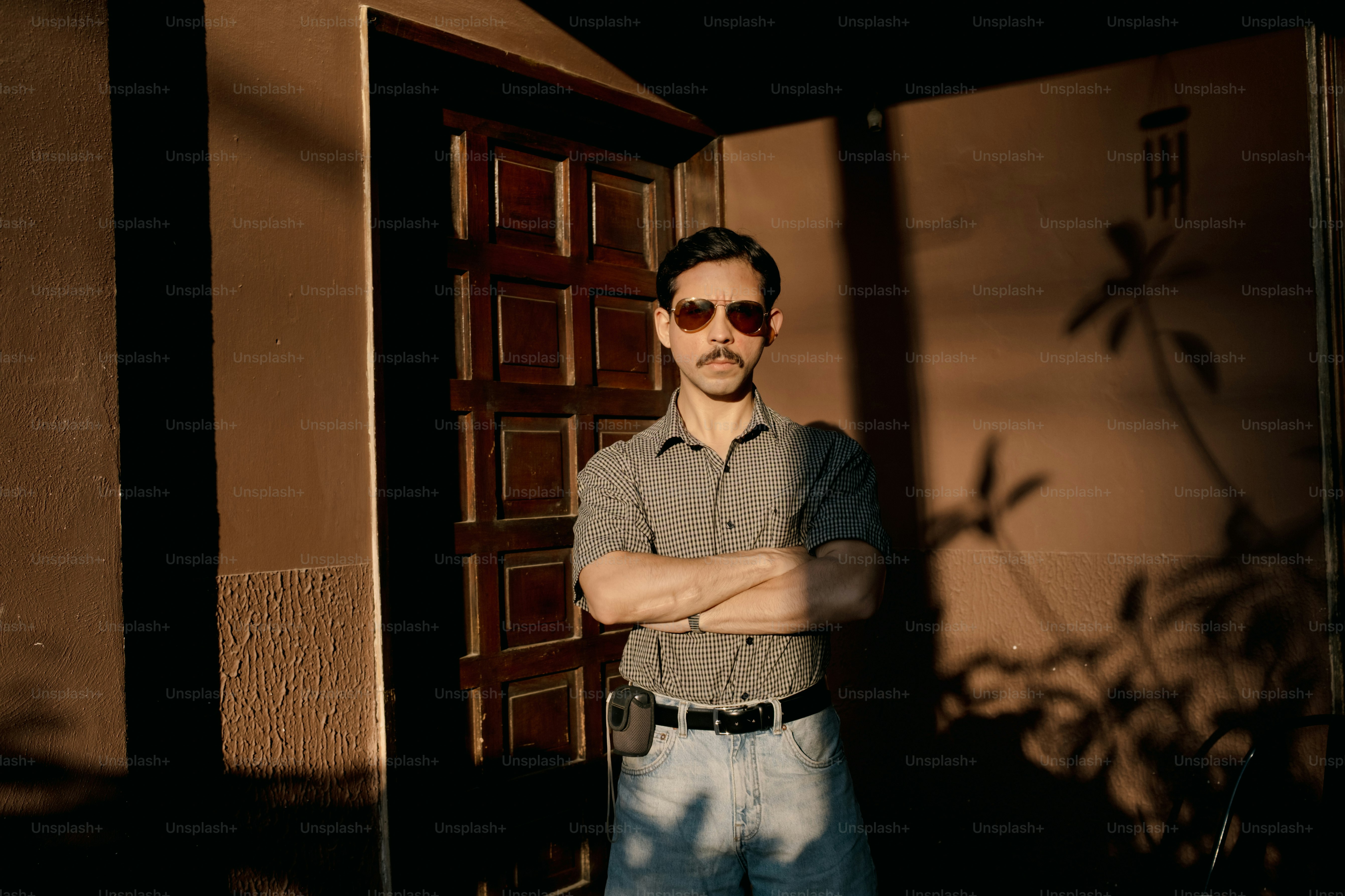 Man with sunglasses and mustache stands with arms crossed