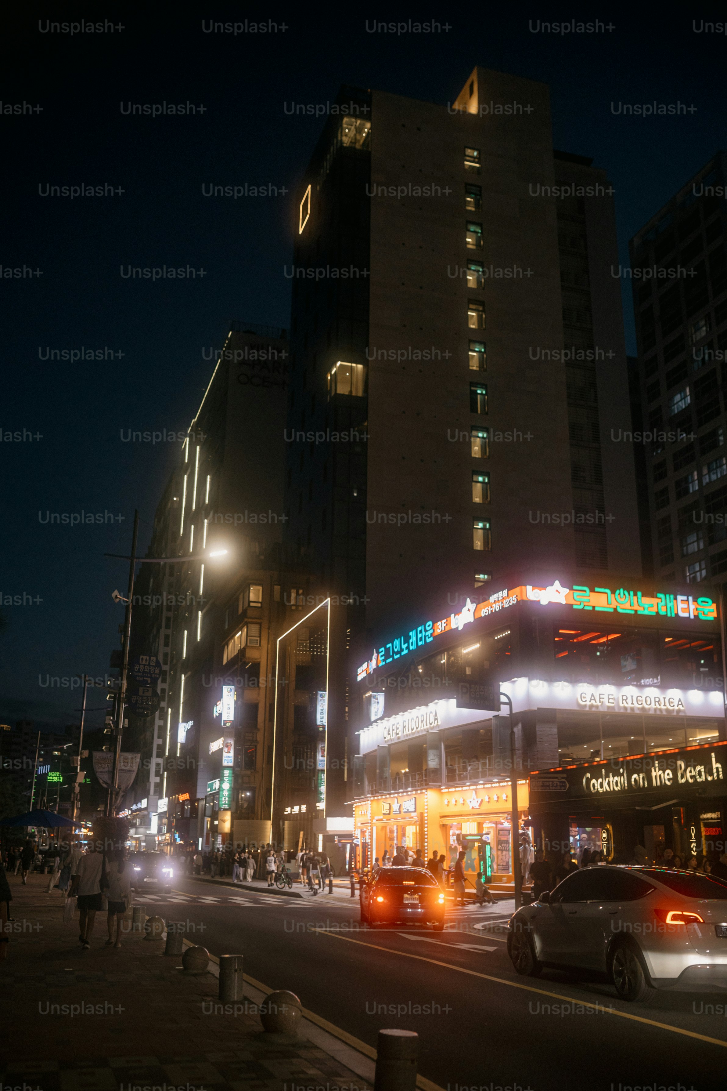 Jalan kota di malam hari dengan bangunan dan mobil yang diterangi.