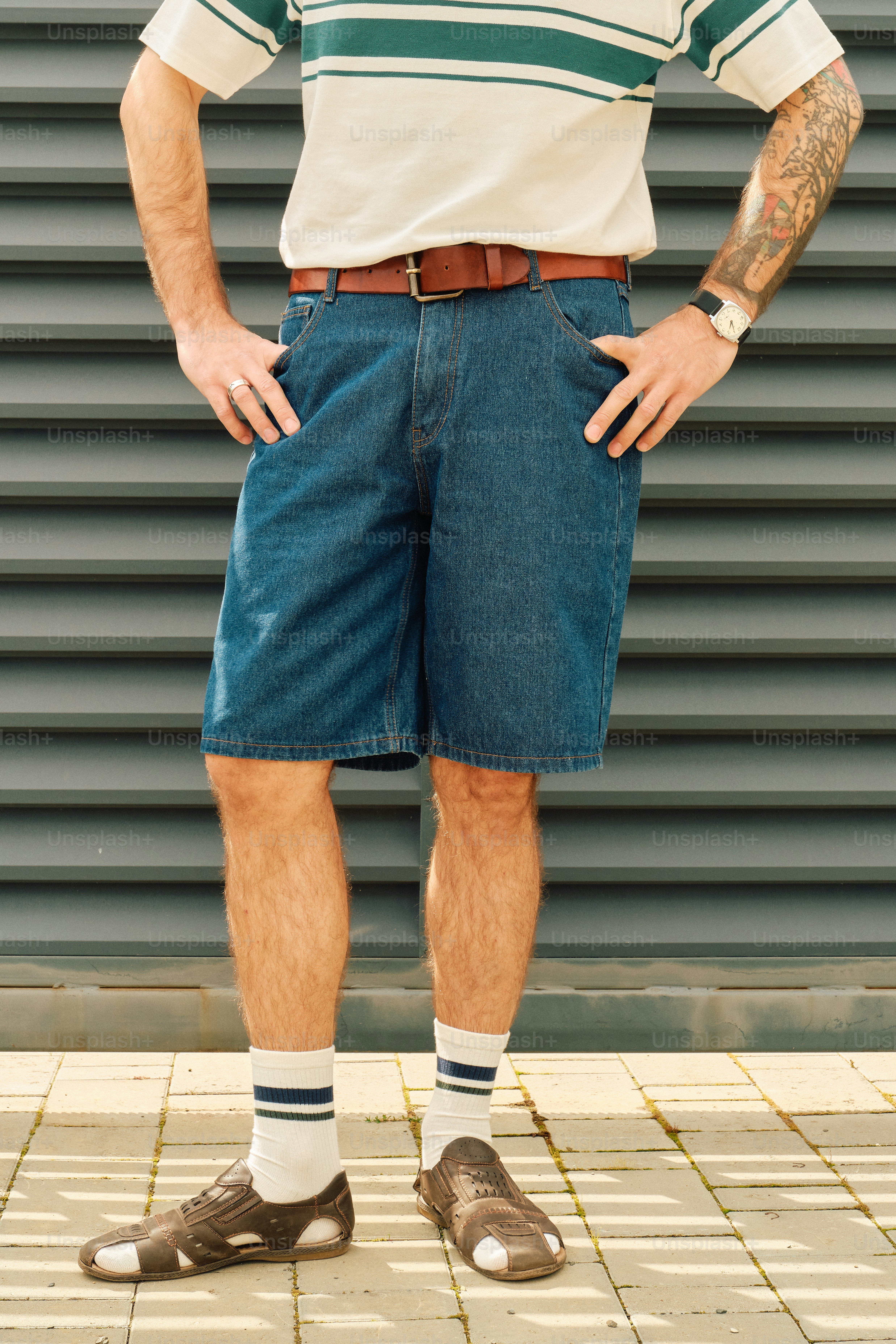 Un homme en short en jean et chemise rayée se tient contre le mur.