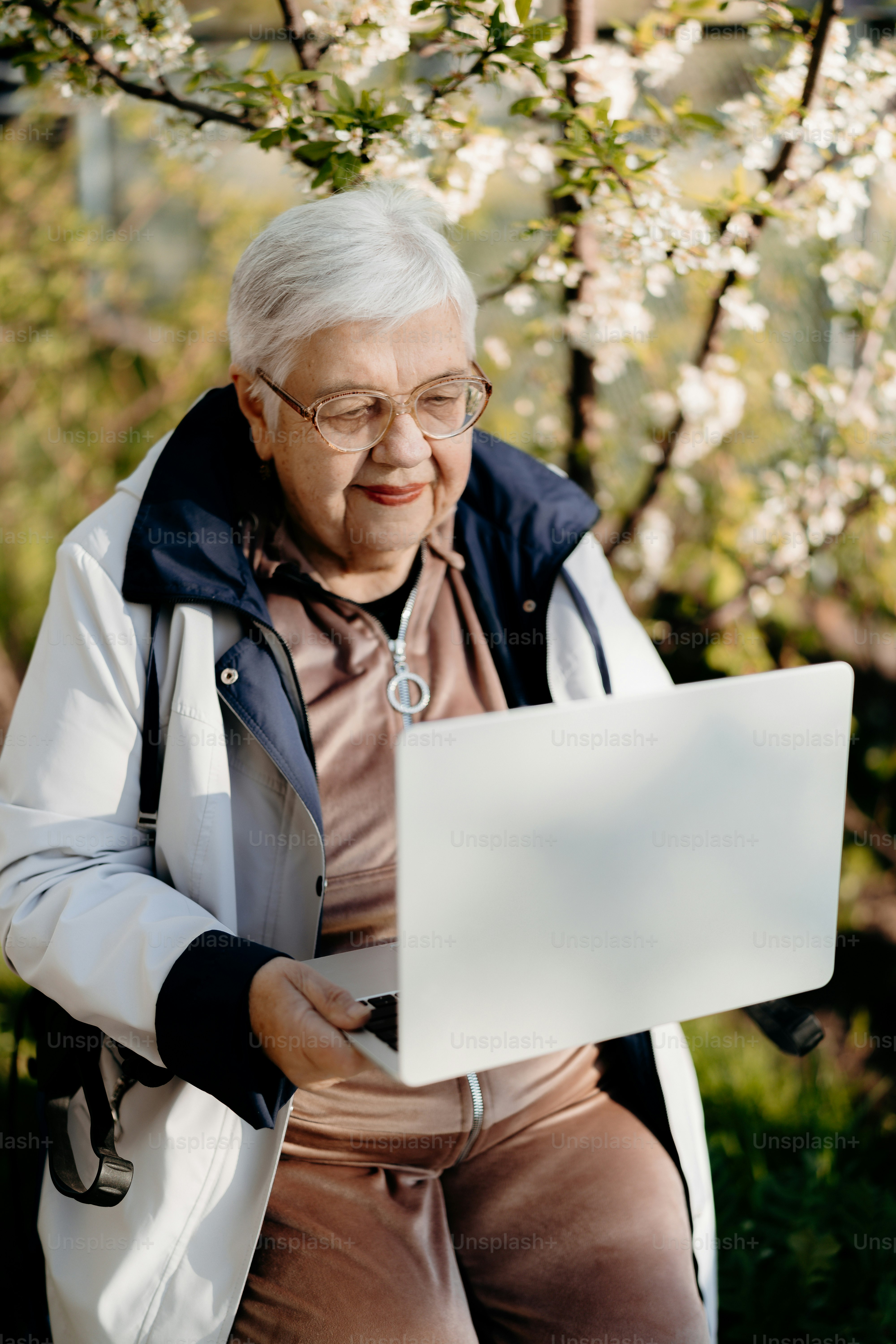 Ältere Frau mit einem Laptop draußen und einem blühenden Baum dahinter