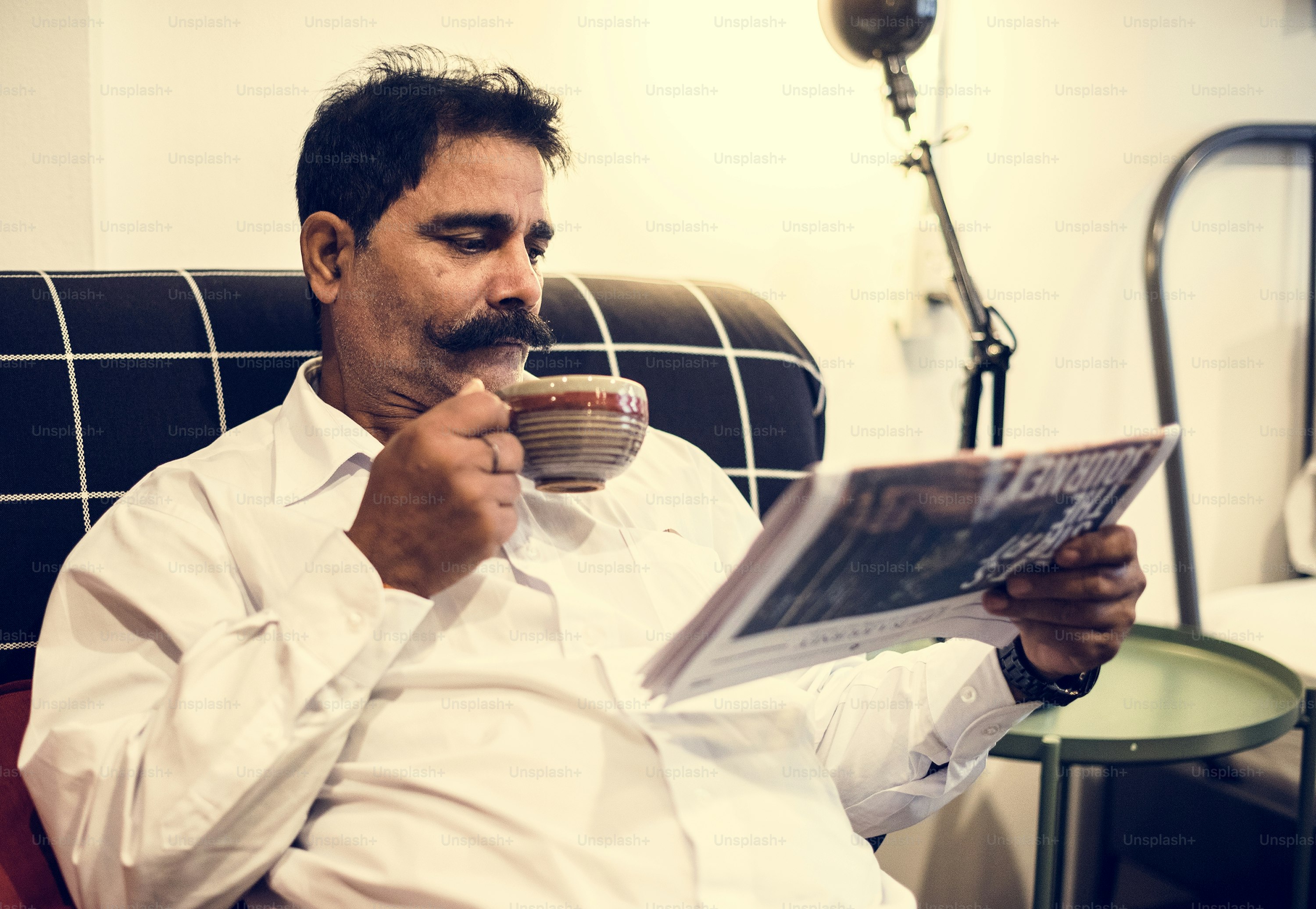 A man with a mustache drinking coffee and reading a newspaper on a sofa