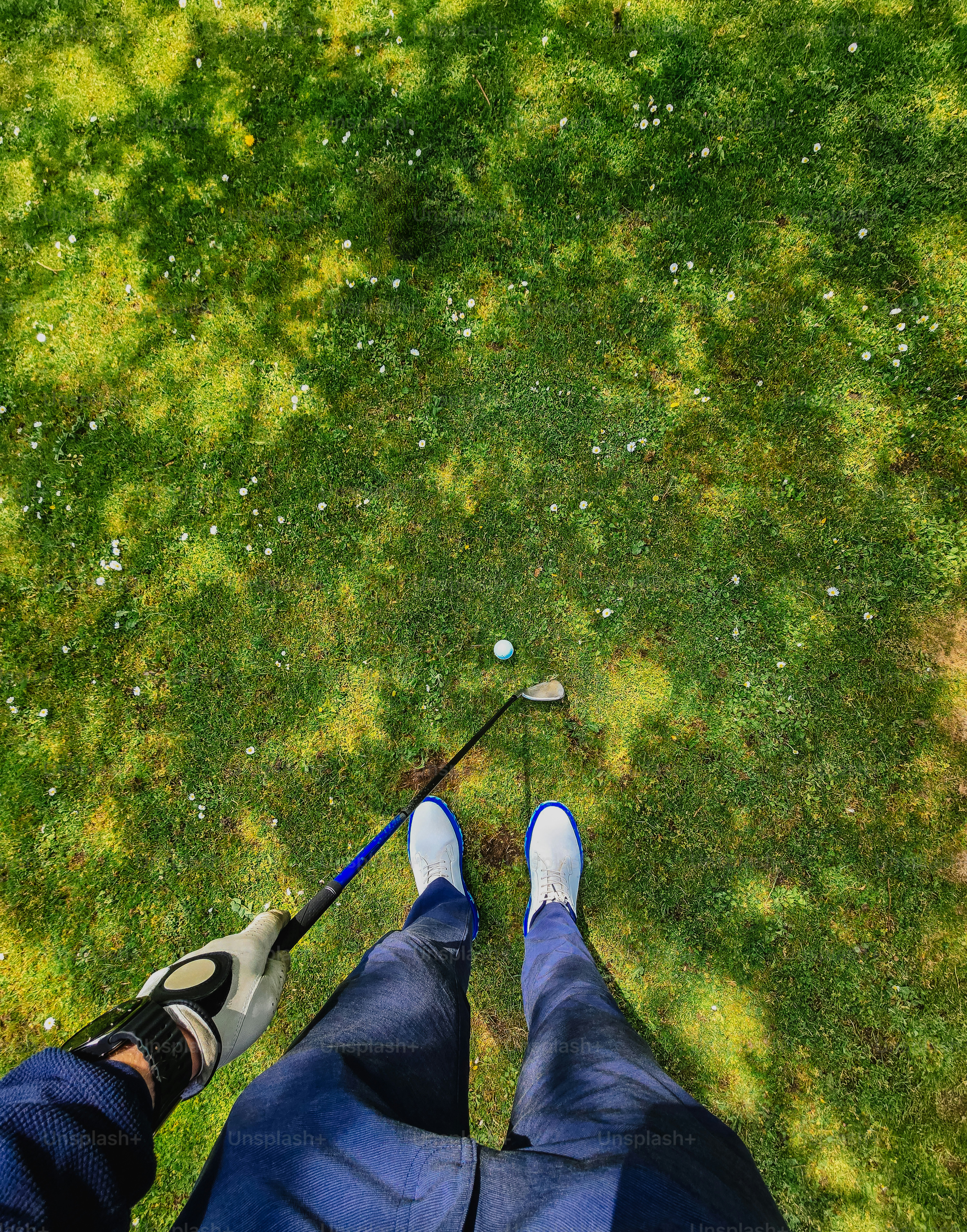 Golfer's legs, club, and ball on green grass with shadows.