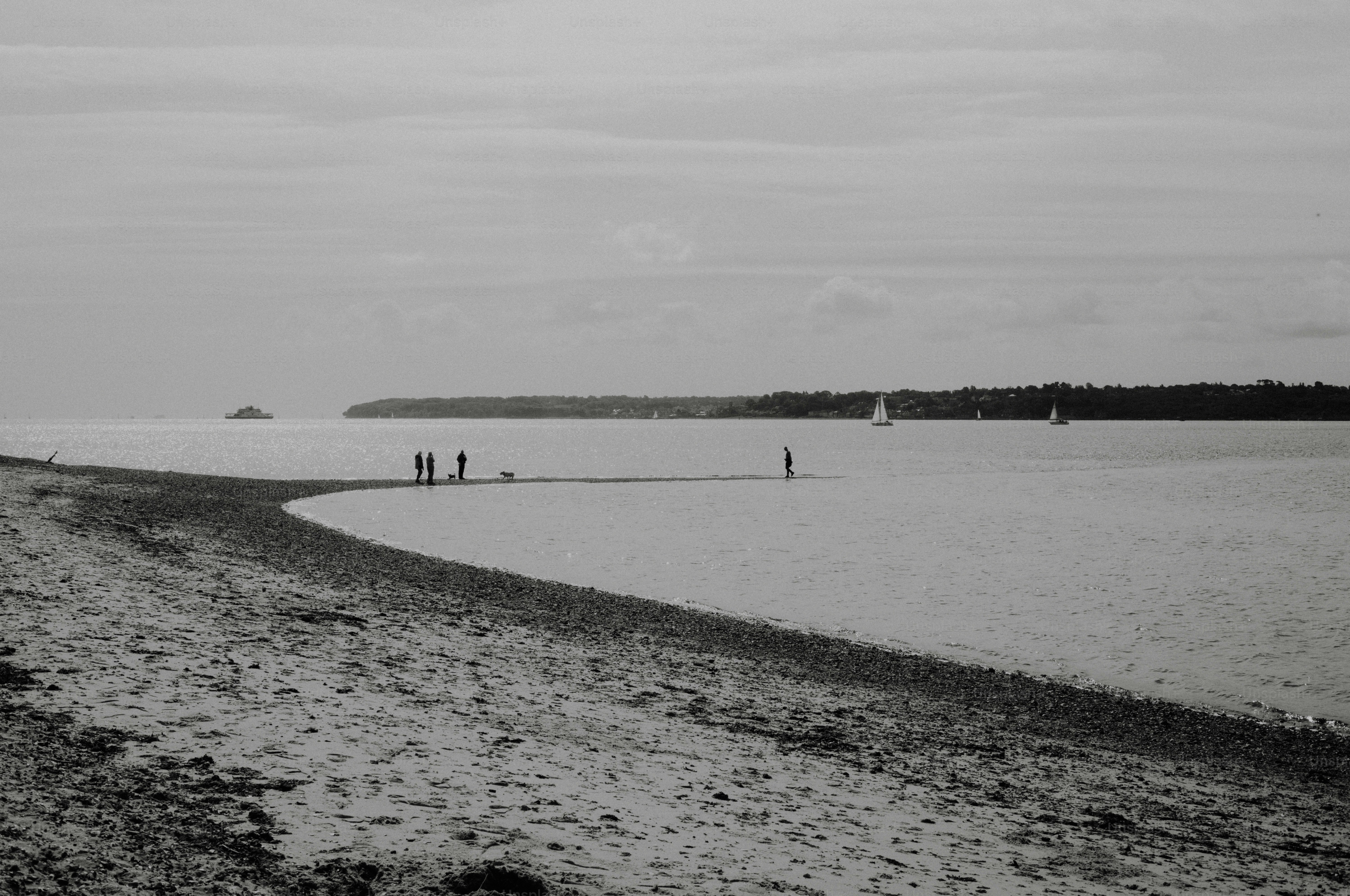 People walking on a sandy beach with sailboats in distance