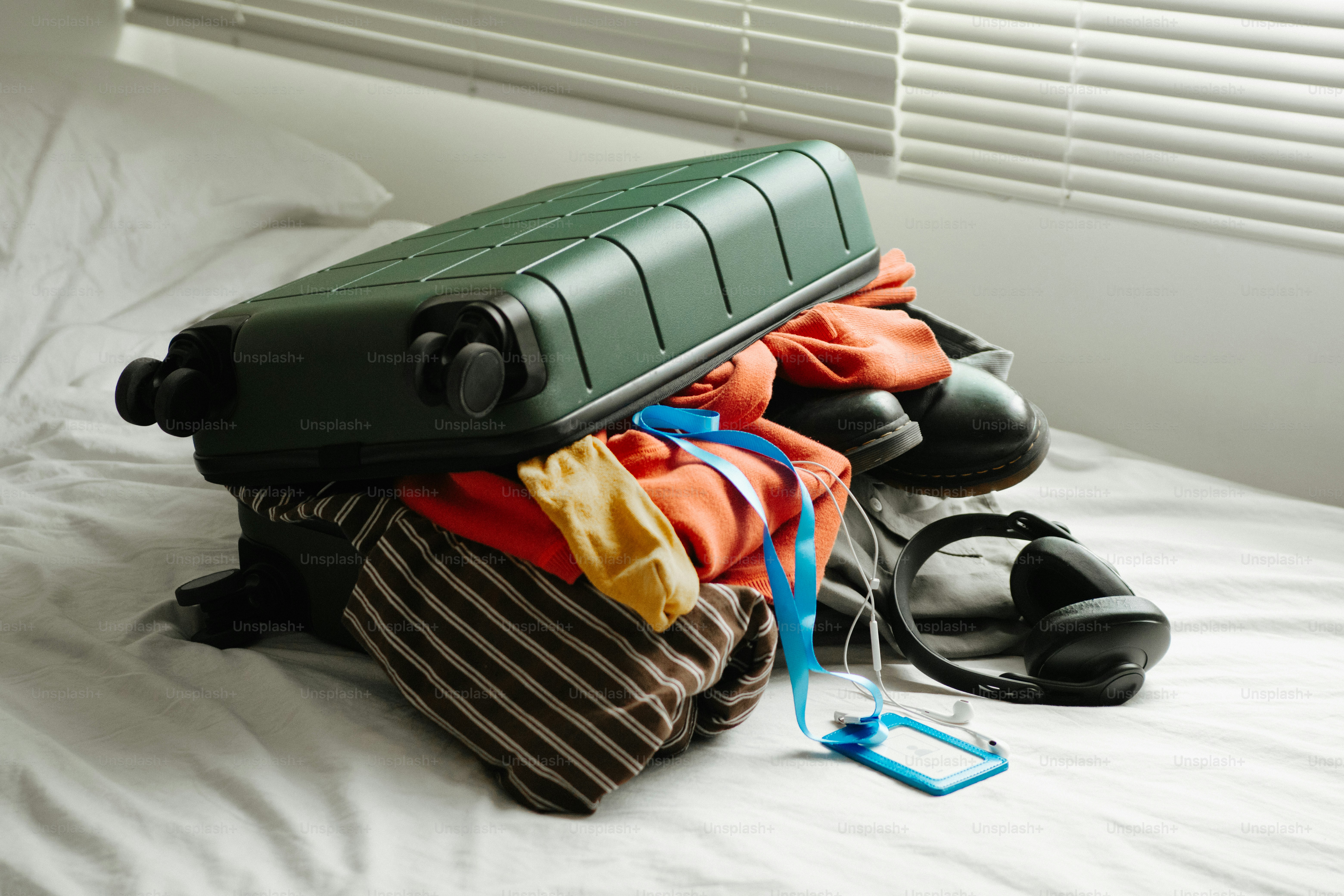 Green suitcase overflowing with clothes on a white bed.