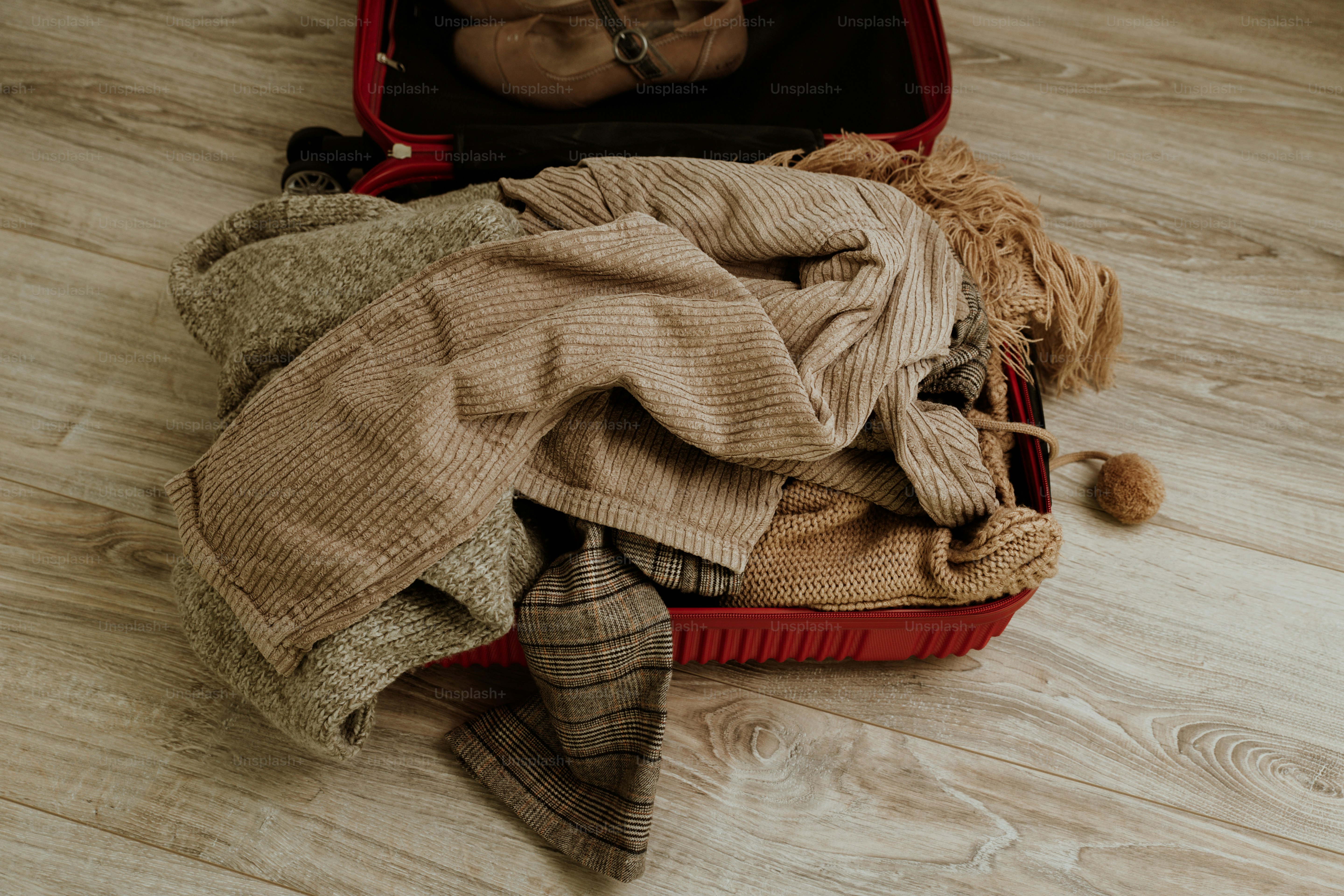 Une valise rouge ouverte débordante de vêtements beiges et marron.