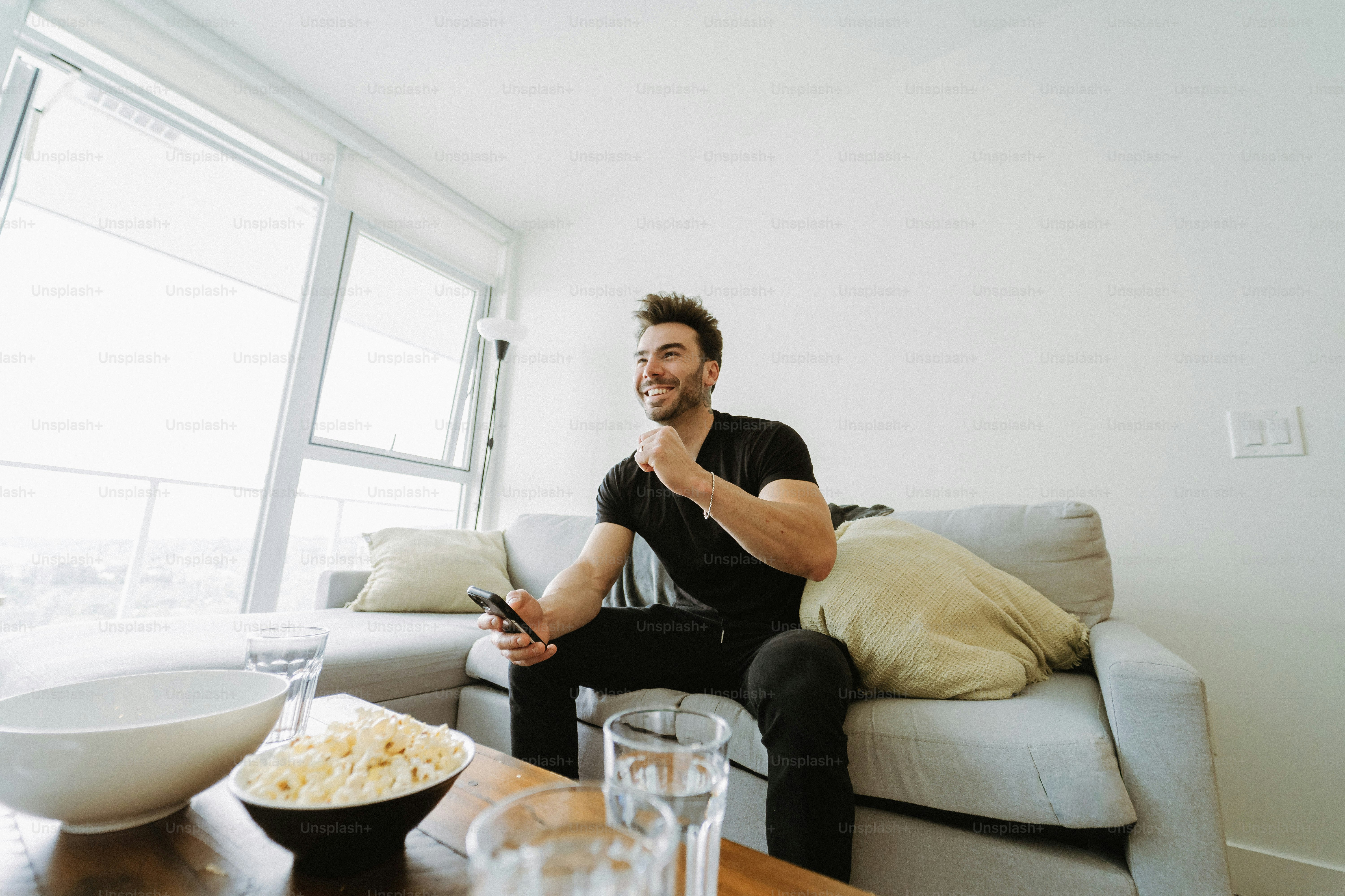Pria tersenyum menonton televisi di sofa dengan popcorn.