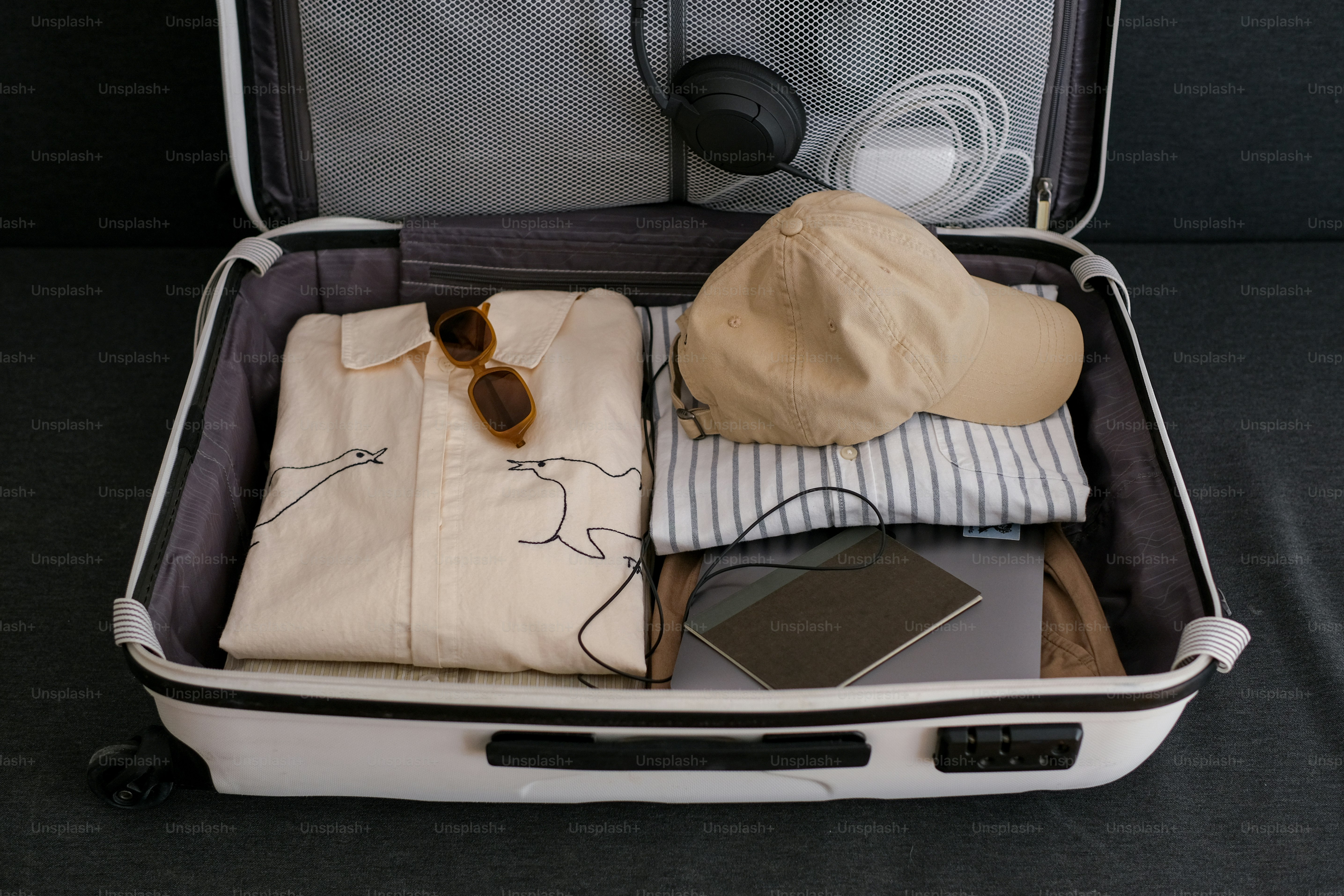 White suitcase packed with travel clothes, hat, and electronics.